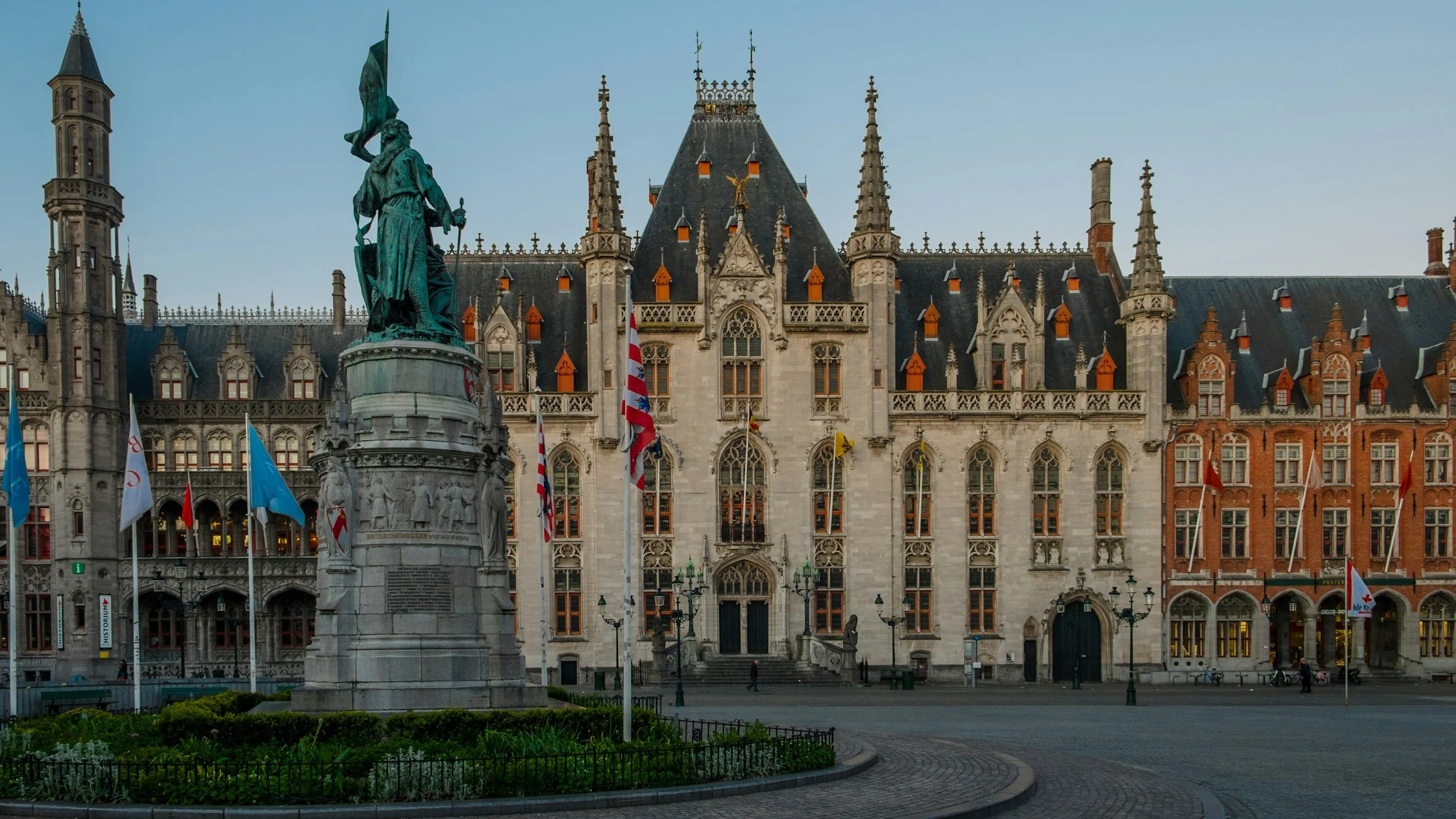 View of a historic building with gothic architecture and a large statue in front, surrounded by flags in a town square.