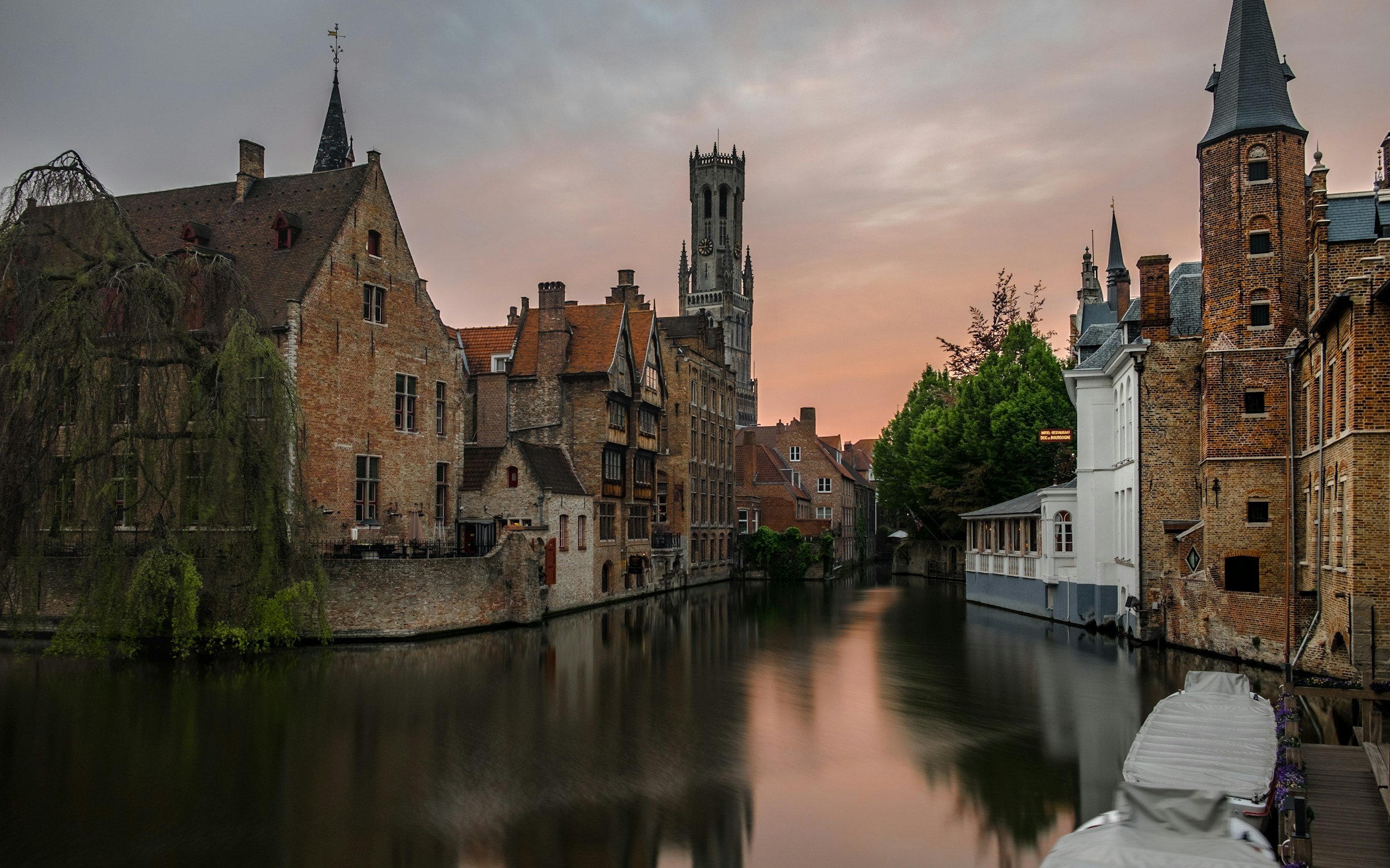 A historic European cityscape with old brick buildings along a canal at sunset, featuring a tall tower with a clock, lush green trees, and a calm water reflection.