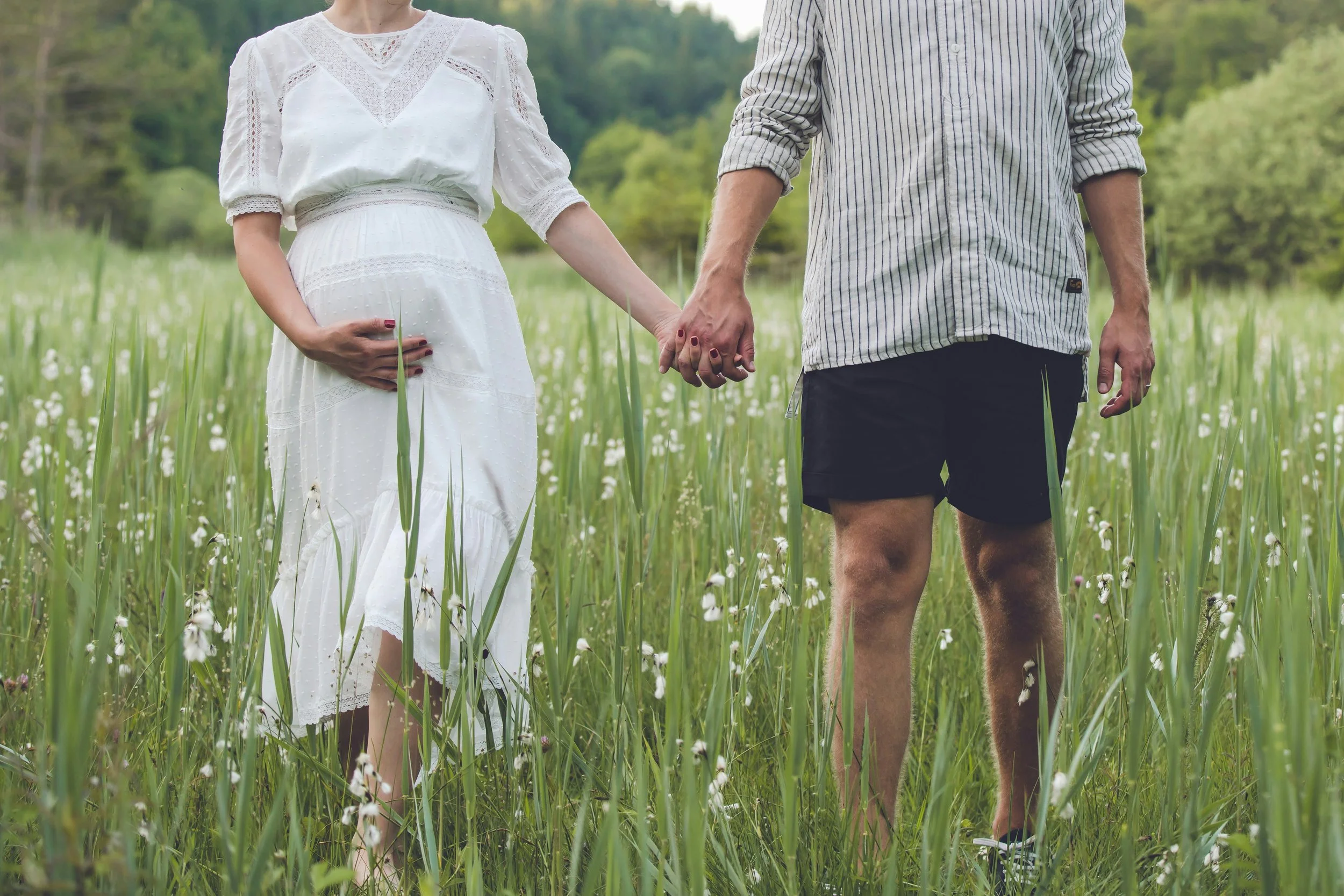 A couple hold hands in a meadow as they go for a walk. The woman is holding her pregnancy bump.