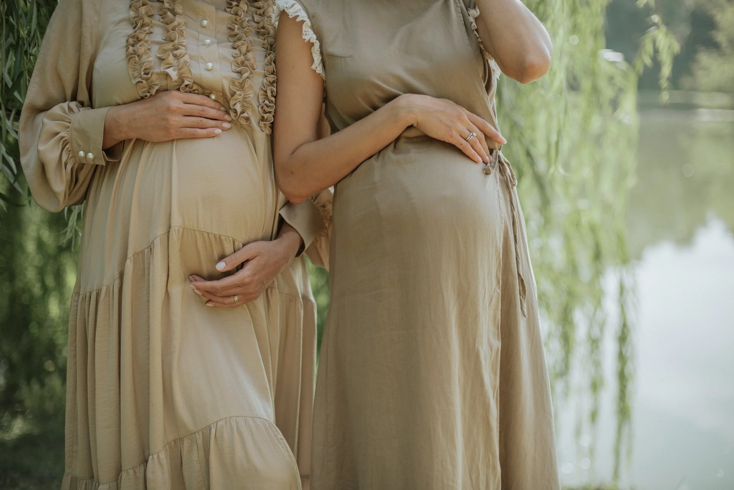 Two pregnant women stand in front of a lake and willow tree holding their baby bumps.