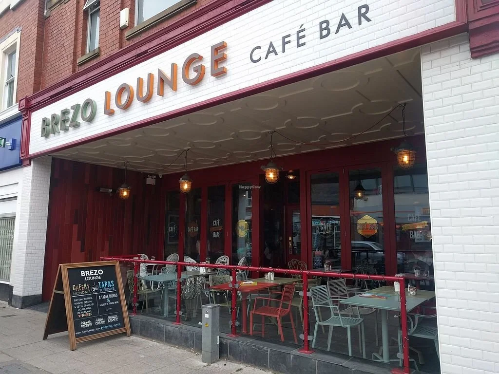 Exterior view of Brezo Lounge Café Bar showing a seating area outside with tables and chairs, red railing, and a blackboard menu sign.