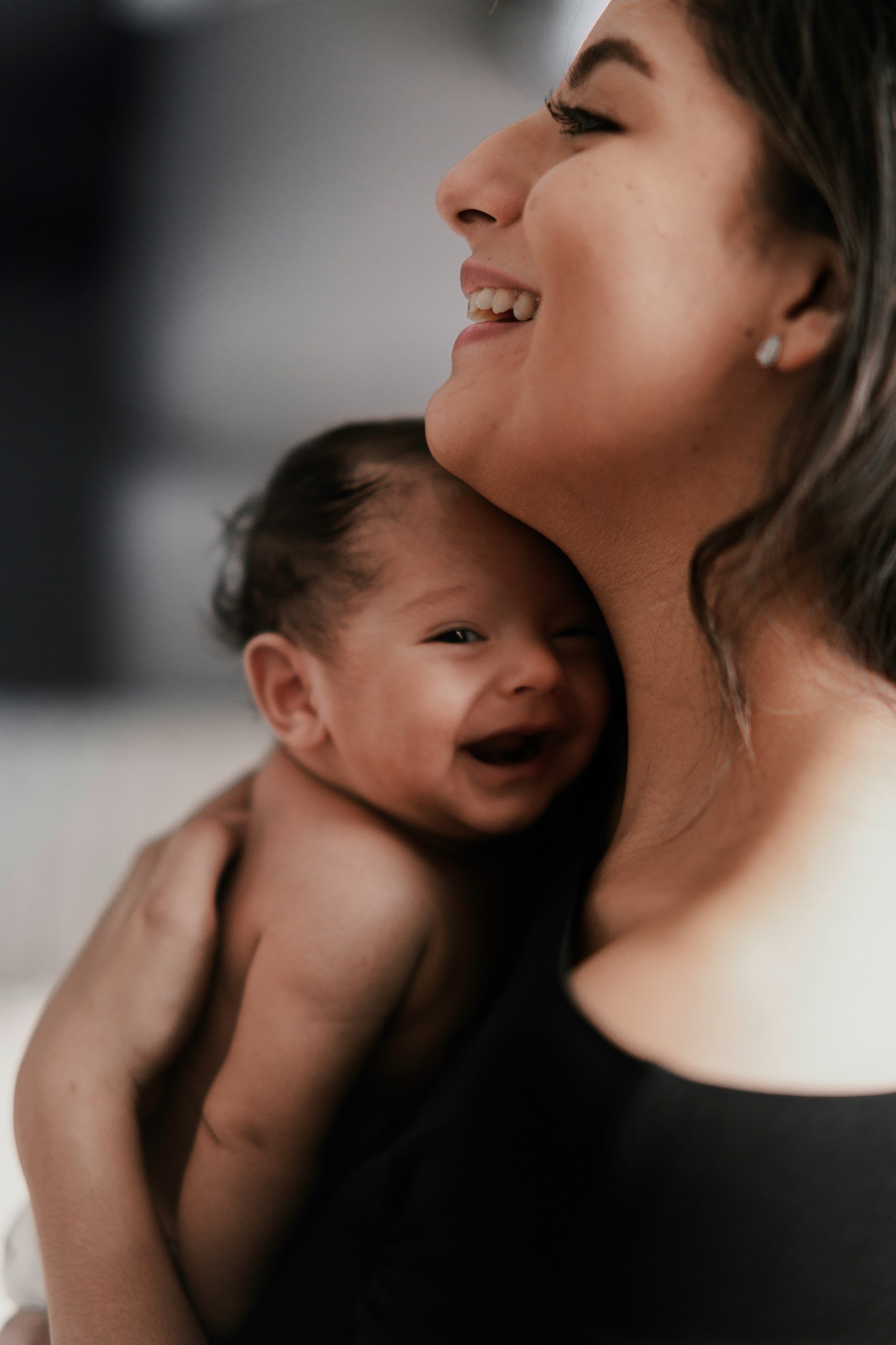 A close-up of a woman with long dark hair and a baby boy with short dark hair, both smiling warmly. The woman is looking to the side, and the boy is looking at the camera.