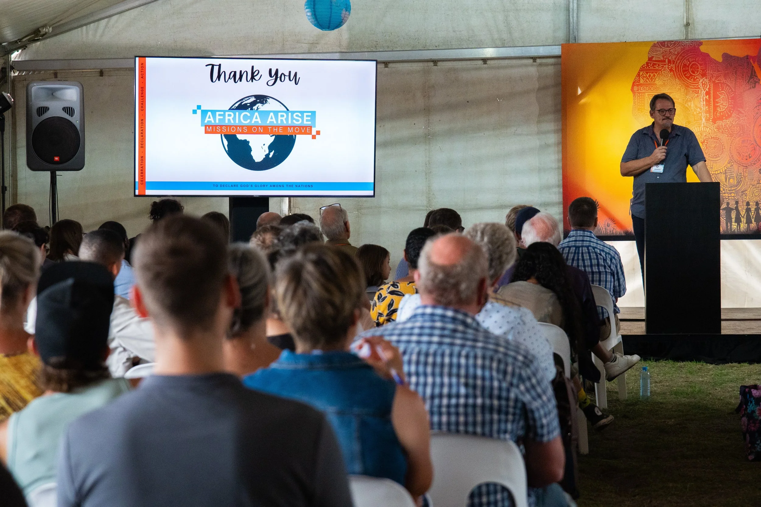 A man standing behind a podium speaking at a conference, with a large screen displaying a thank you message and a logo that reads 'Africa Arise Missions on the Move' to the left, and an audience seated facing the stage within a large tent. conference