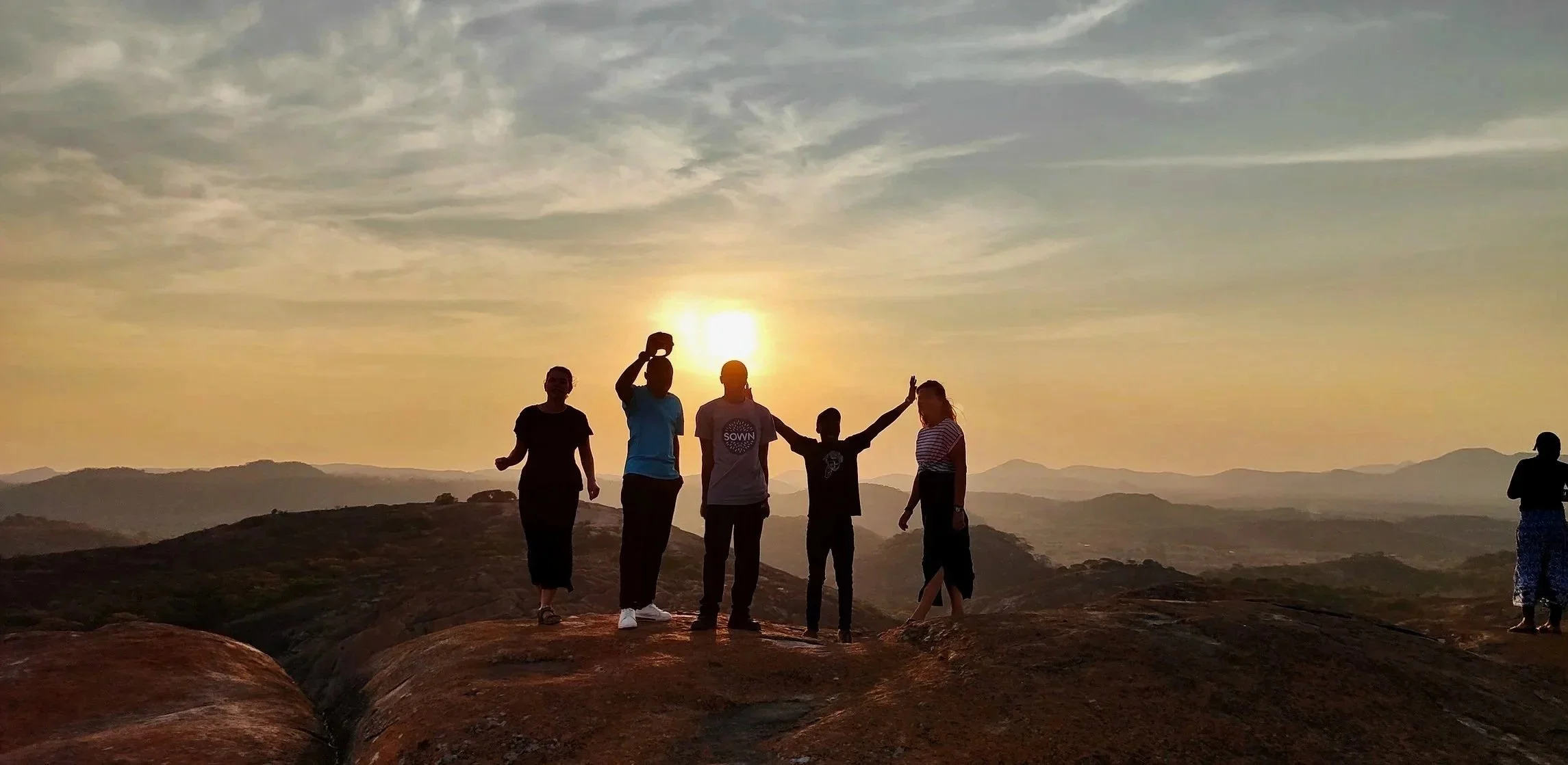 Group of people silhouetted against a sunset atop a rocky hill, with mountains in the distance.