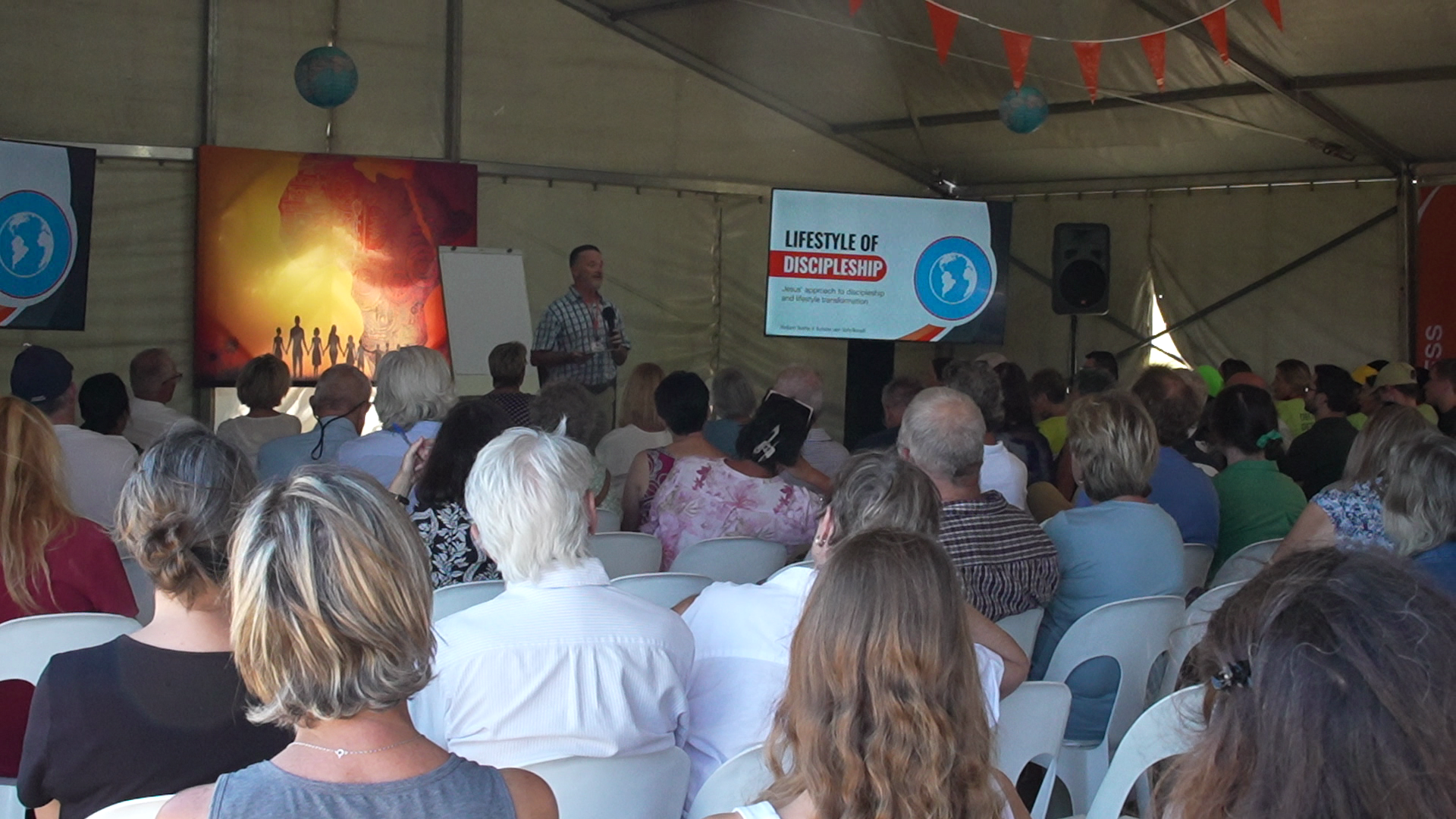 An indoor event with a speaker presenting on the importance of discipleship, titled 'Lifestyle of Discipleship,' with an audience of diverse adults seated and listening attentively.