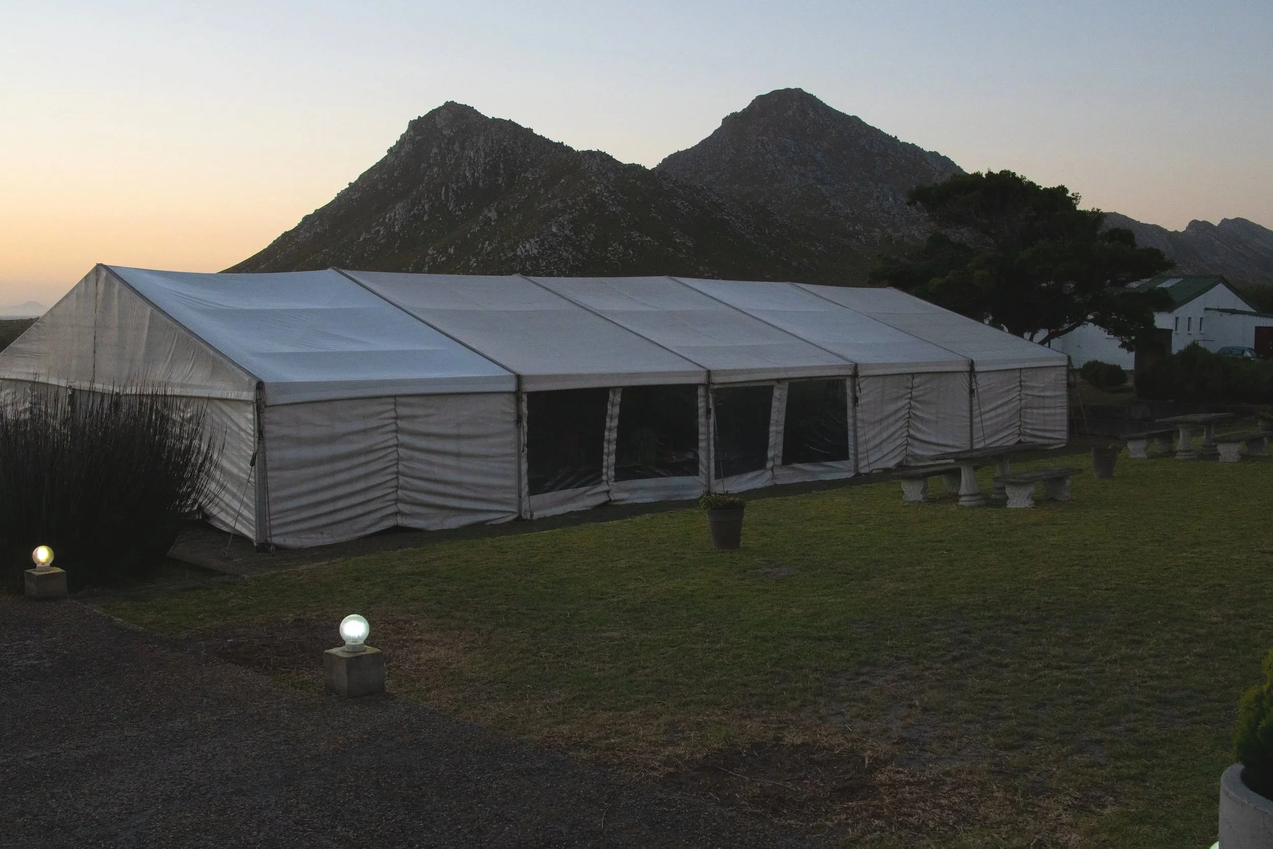 A large white event tent with transparent windows is set up on a grassy area at sunset, with mountains and a single tree in the background.