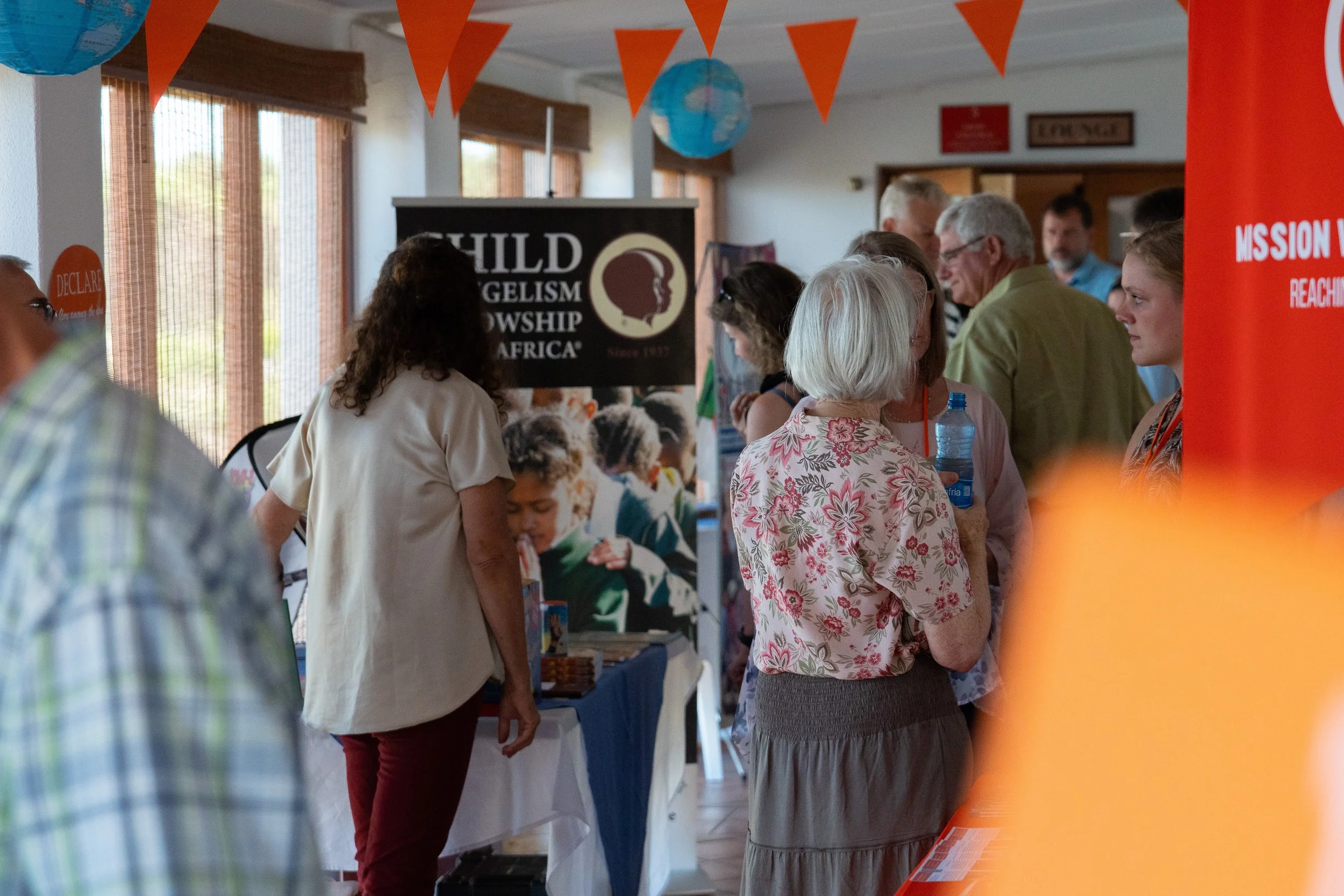 A group of people inside a room at a community event, browsing booths with banners and informational material. Orange triangular flags hang from the ceiling and there are windows with blinds along the wall. Exhibitors taking hands