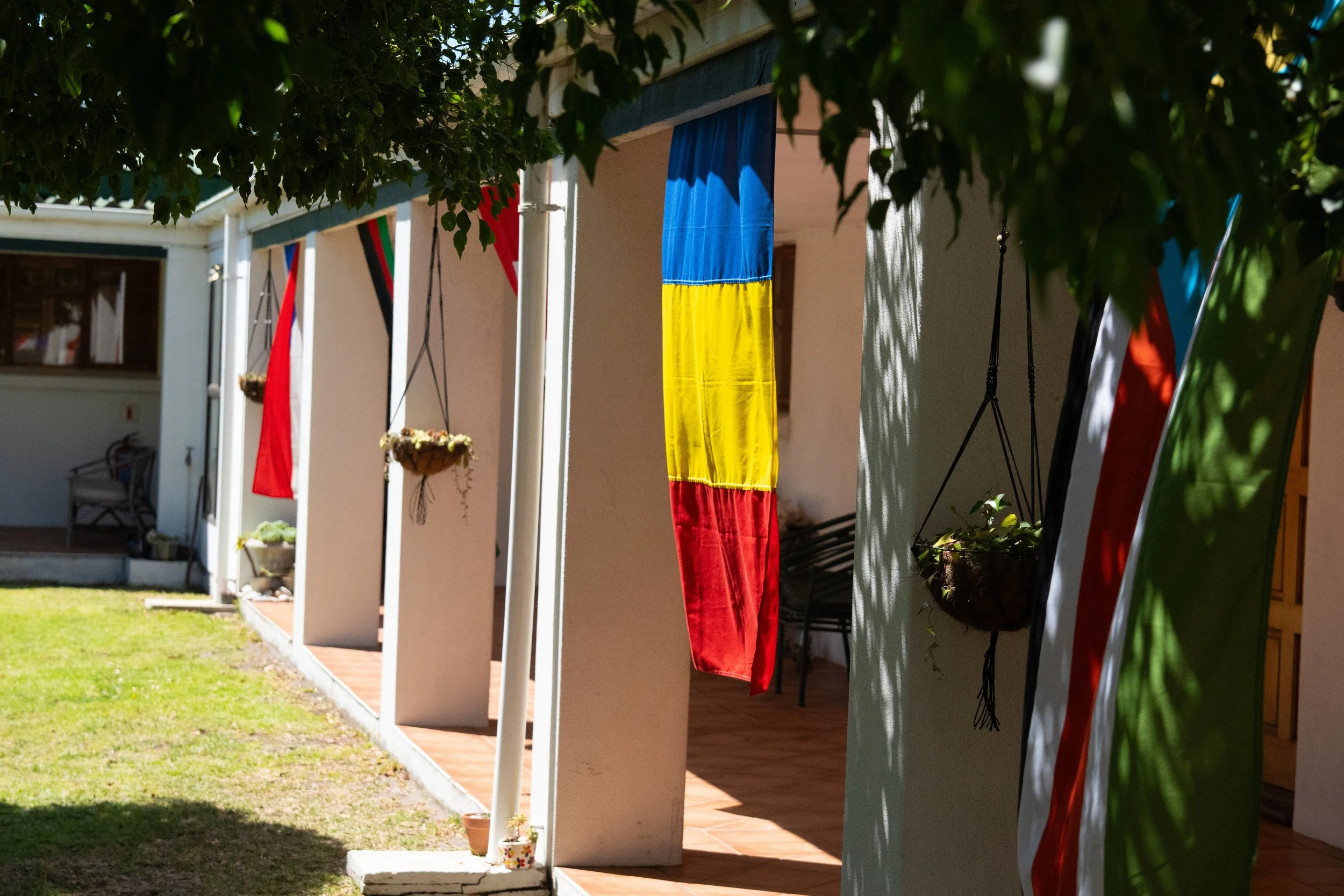 Colorful flags hanging on a porch, with a focus on a flag with blue, yellow, and red vertical stripes, and other red, black, and green flags in the background, along with potted plants and outdoor furniture.