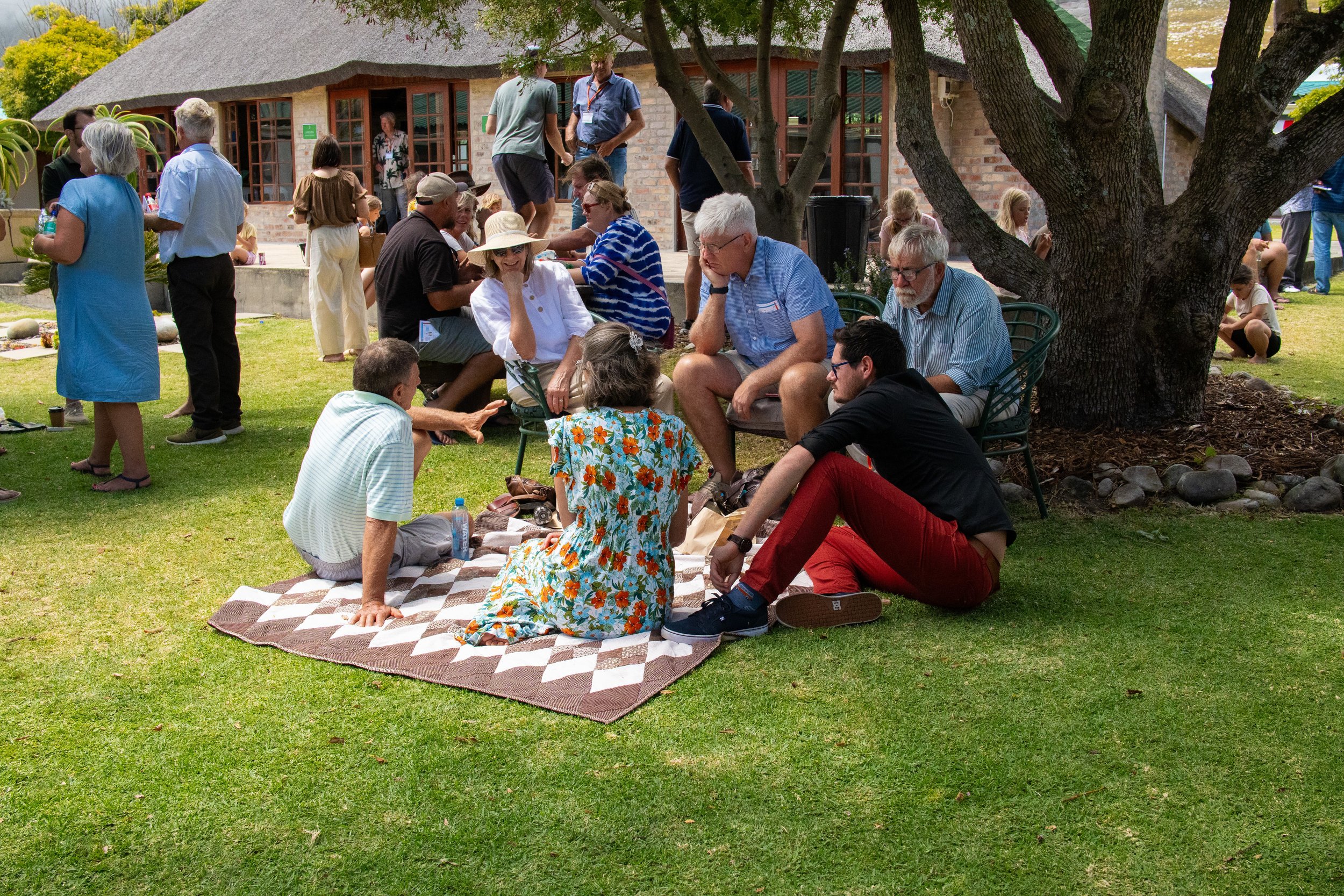 Group of people gathered outdoors on a sunny day, sitting on a blanket and chairs under a tree, with other people standing and walking nearby in front of a brick house with a thatched roof.