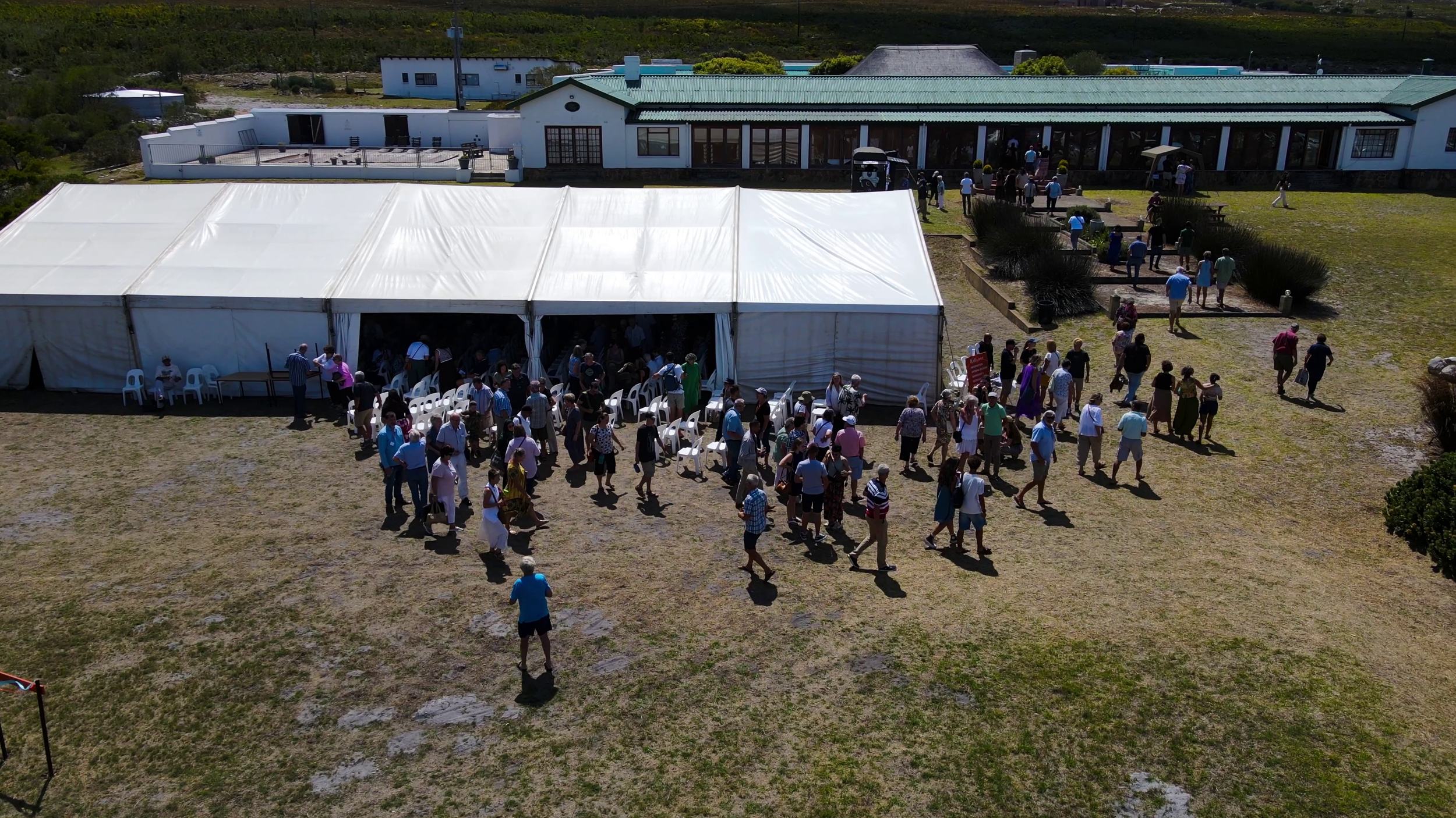 A large outdoor event with many people entering a white event tent. The scene takes place on a grassy area in front of a building with a green roof, and there are trees and additional structures in the background.