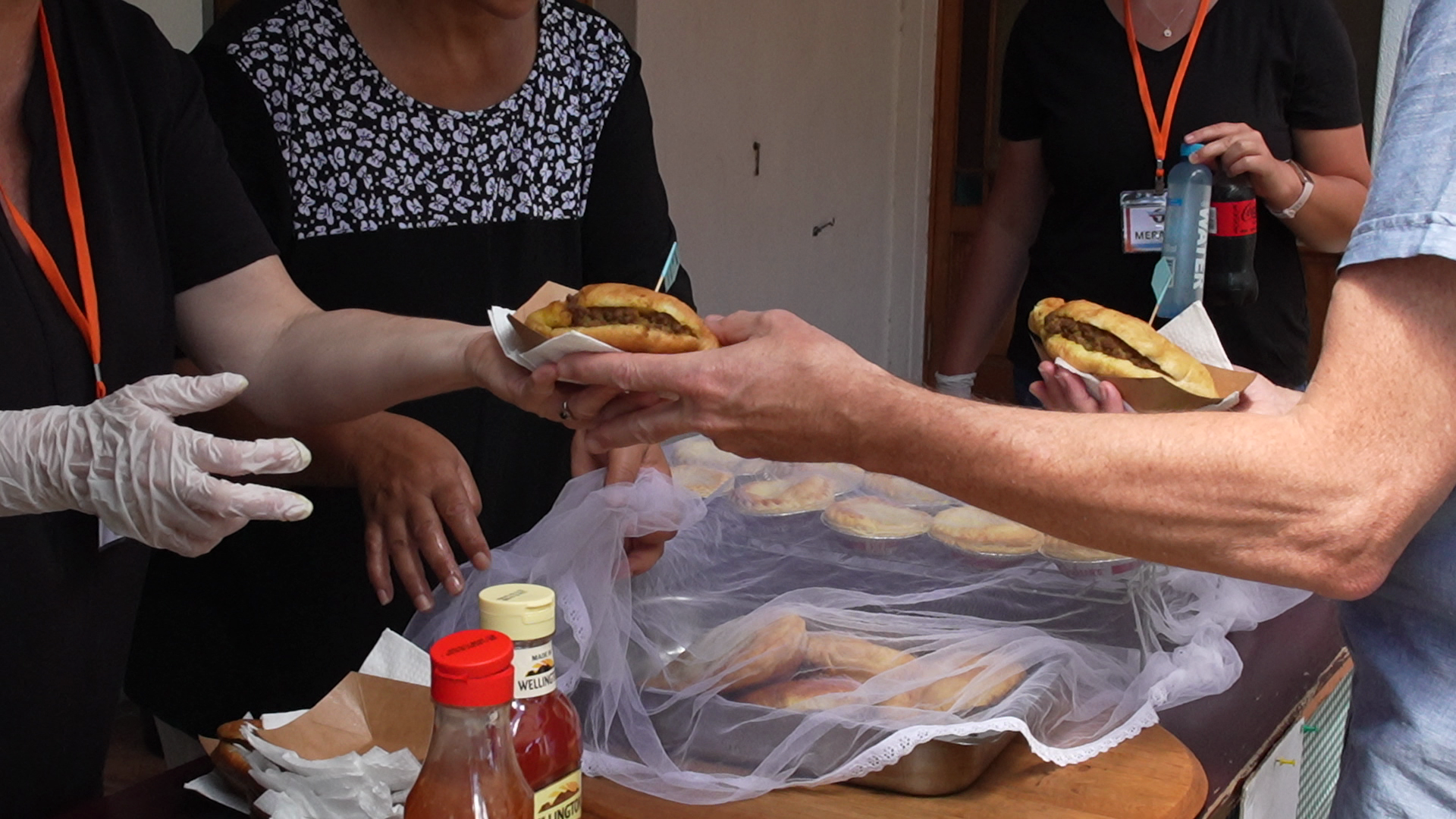 People serving and receiving hot dogs at a community or charity event. Hands passing or holding hot dogs, with condiments and food containers on the table.