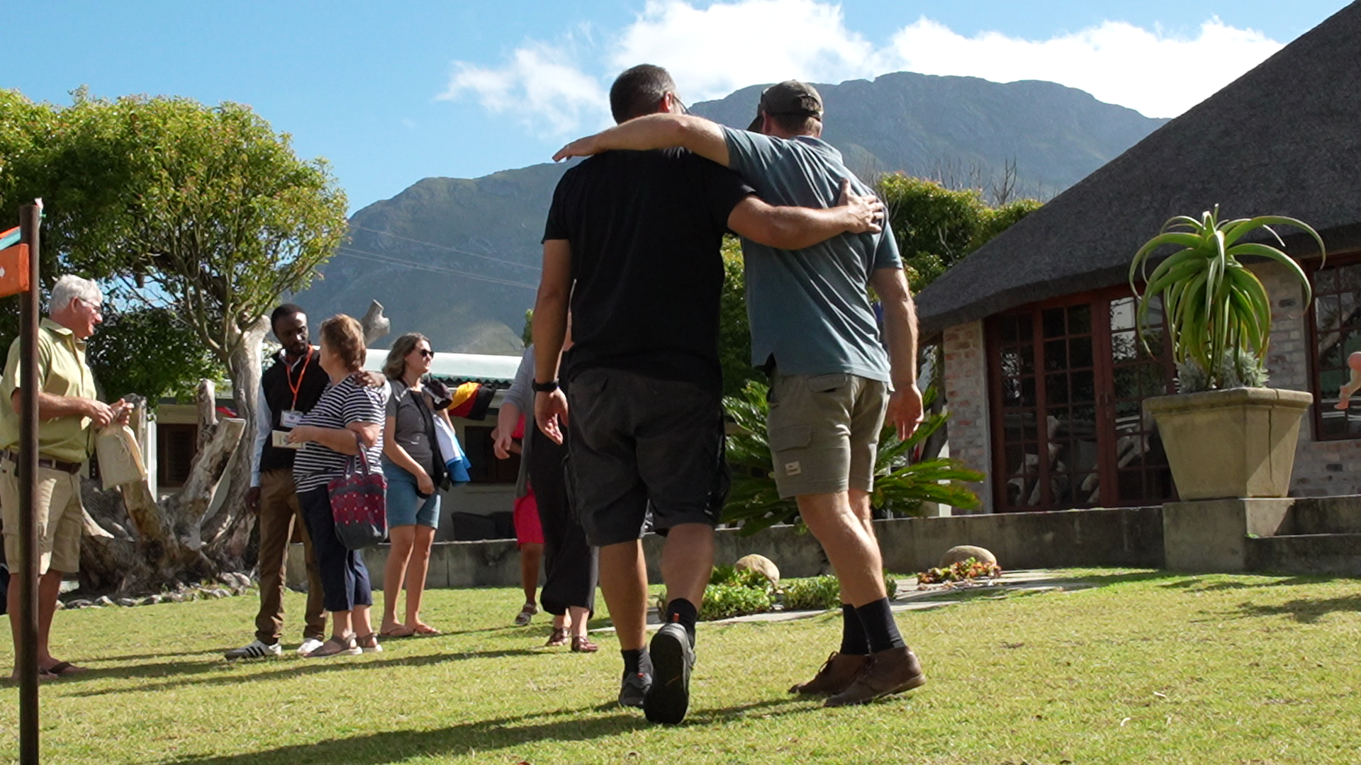 Two men hugging, one with his arm around the other's shoulders, walking on a grassy area with a group of people in the background, trees, a mountain, and a building with large windows.
