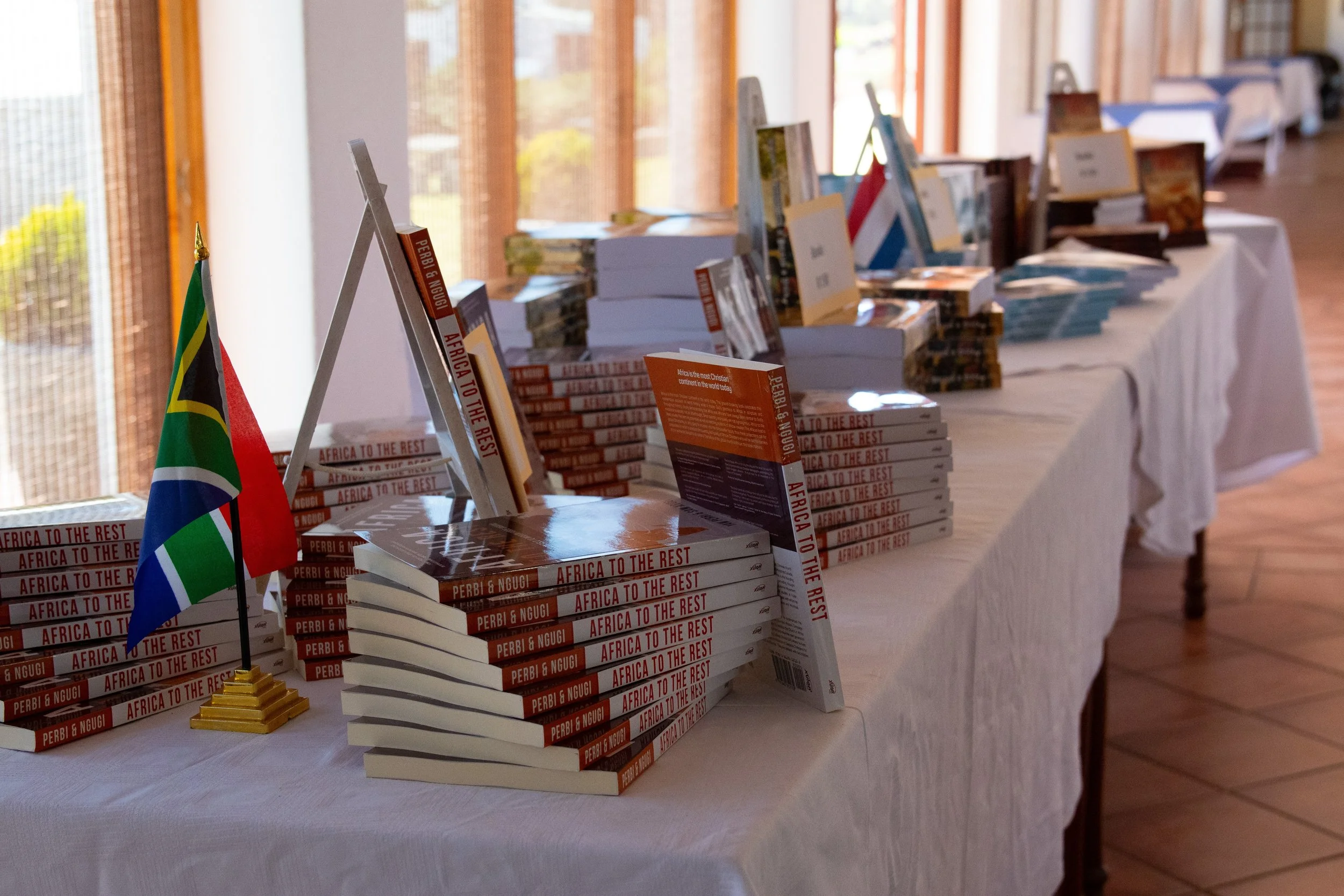 Book display table featuring multiple copies of 'Africa to the Rest' by Perbi & Ngugi, a small South African flag, and various books arranged in stacks in a well-lit room with large windows and wooden flooring.