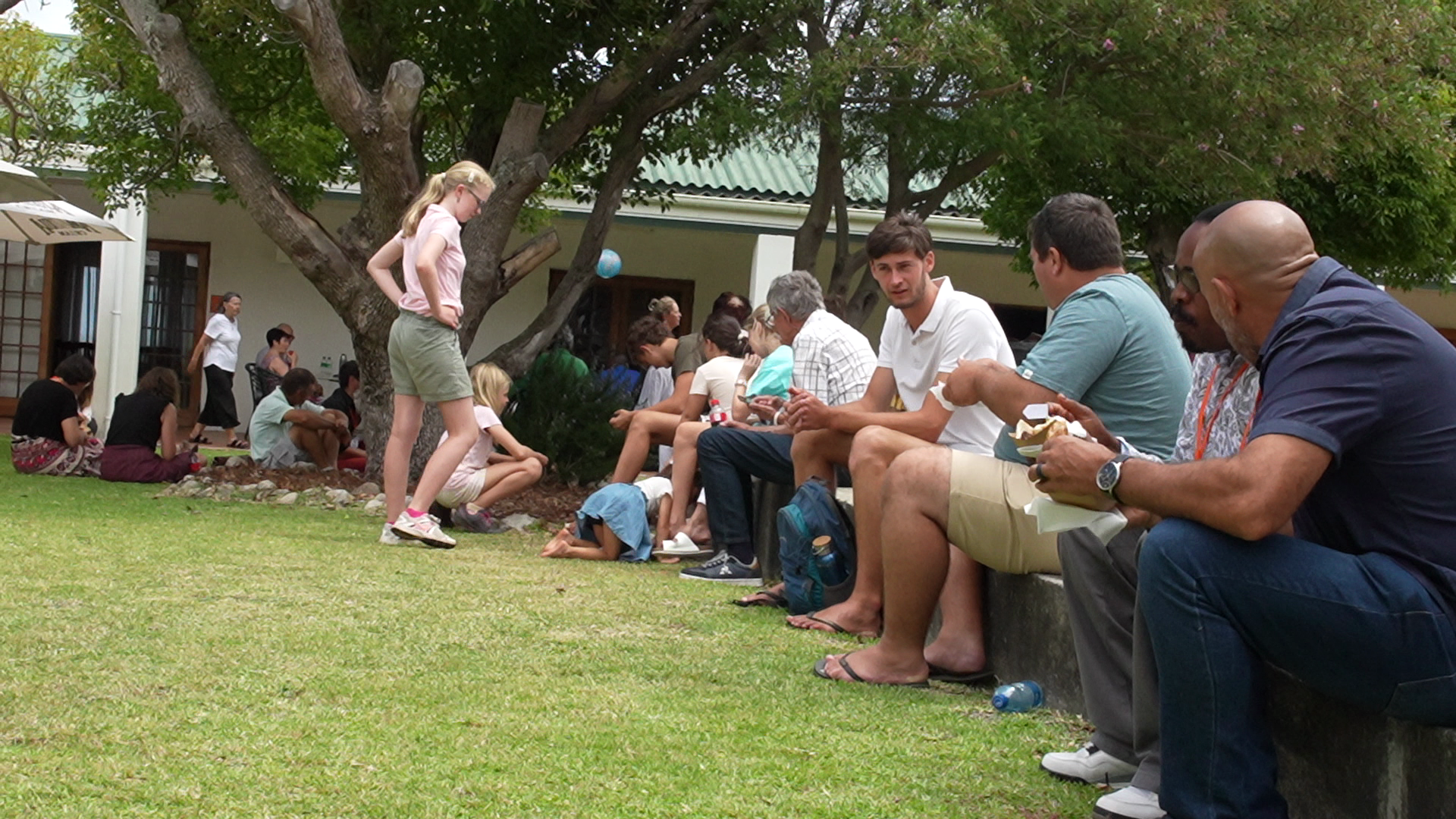 People sitting on a bench and on the ground under trees in a park or garden area, some eating and others chatting or looking at their phones.