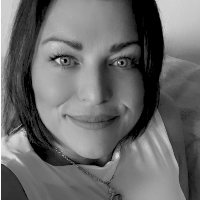 Close-up of a woman with short dark hair, light eyes, and a friendly expression, wearing a white top and a necklace.