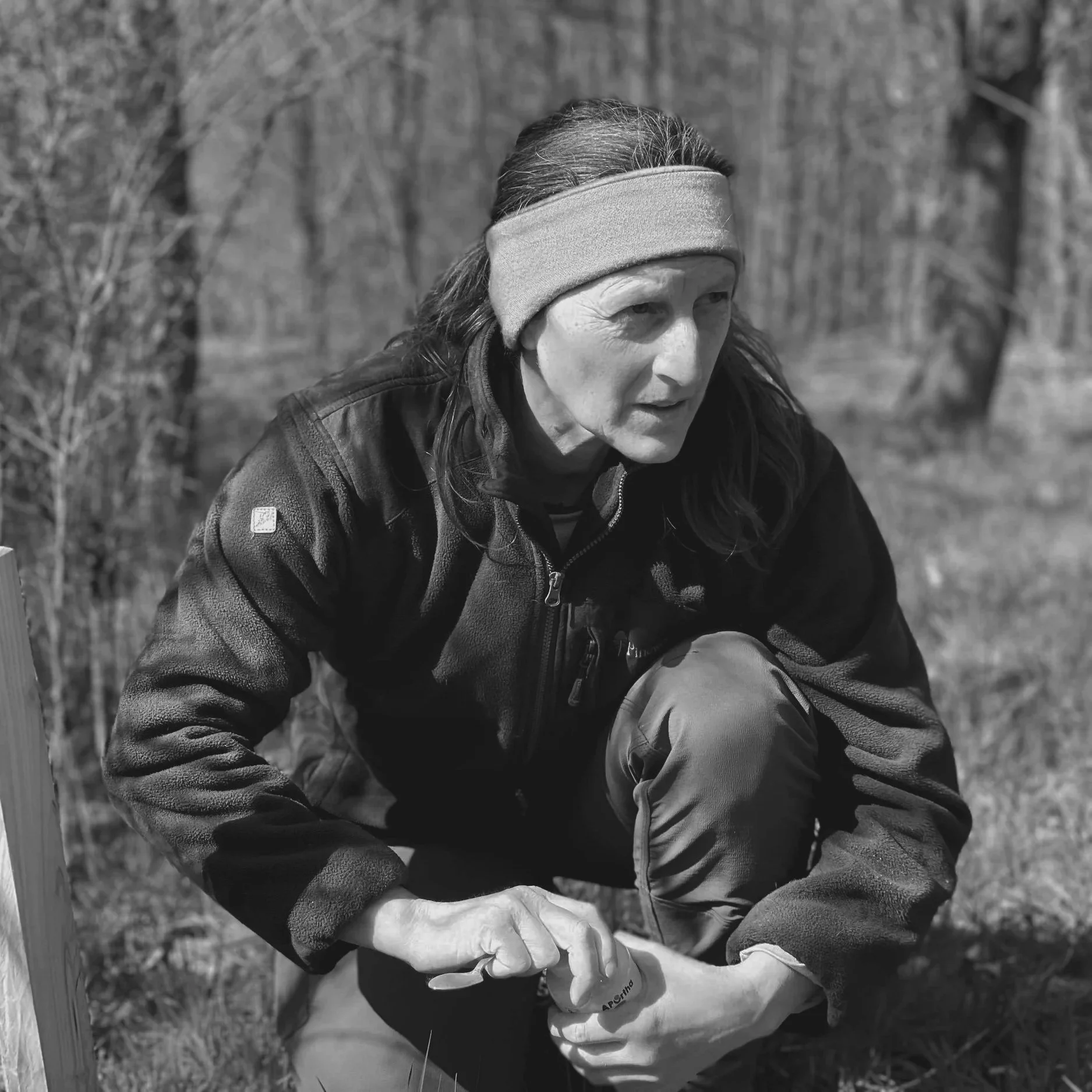 A woman with long hair, wearing a headband, a jacket, and outdoor pants, crouches in a forested area.