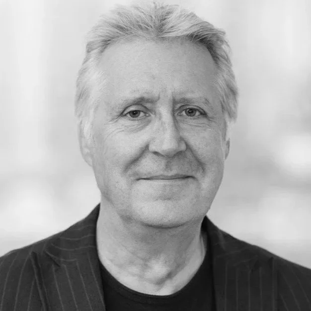Black and white portrait of a middle-aged man with light-colored hair, wearing a dark pinstripe blazer and a dark shirt, smiling softly.