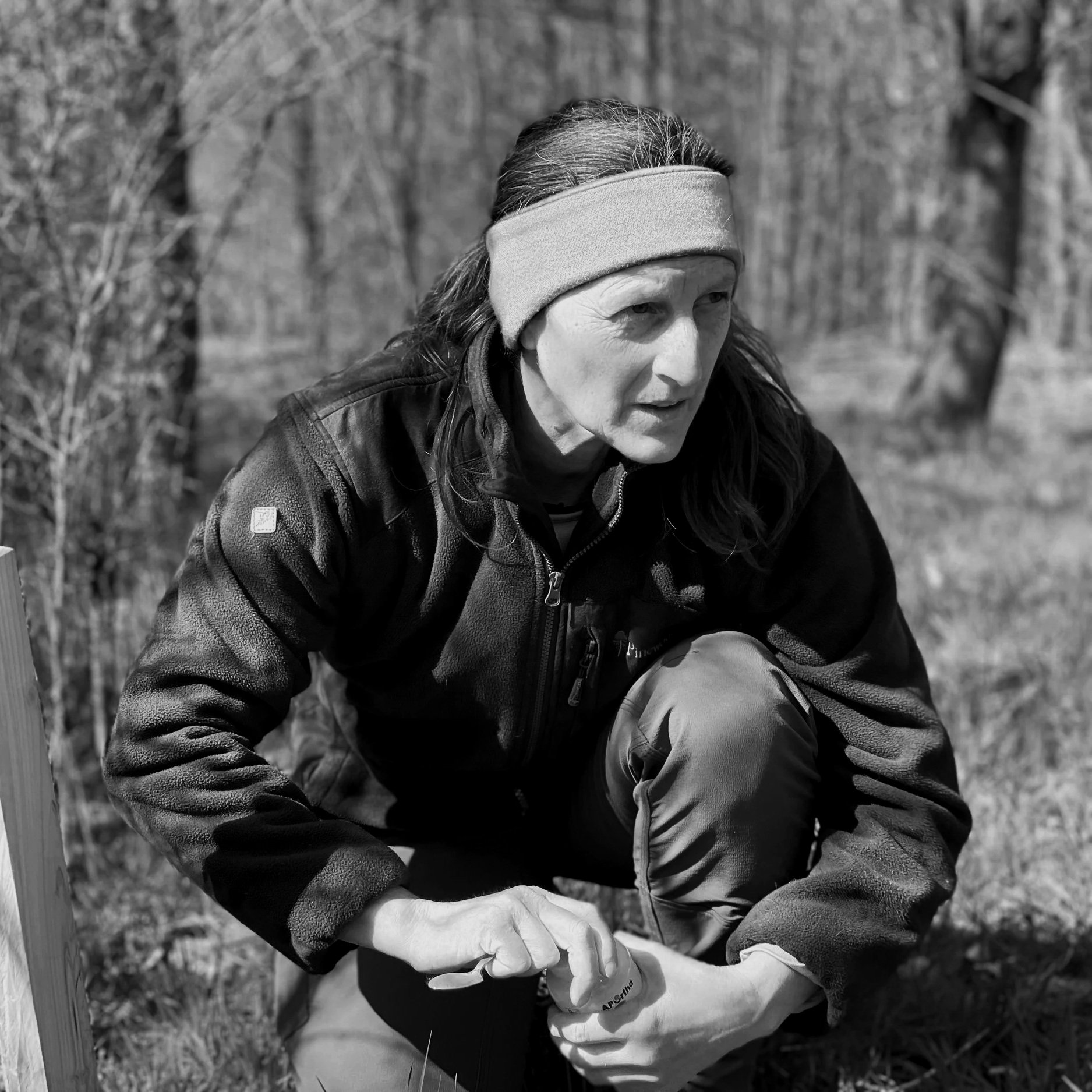 A woman wearing a headband and outdoor jacket crouches near the ground in a wooded area, focused on something in front of her.