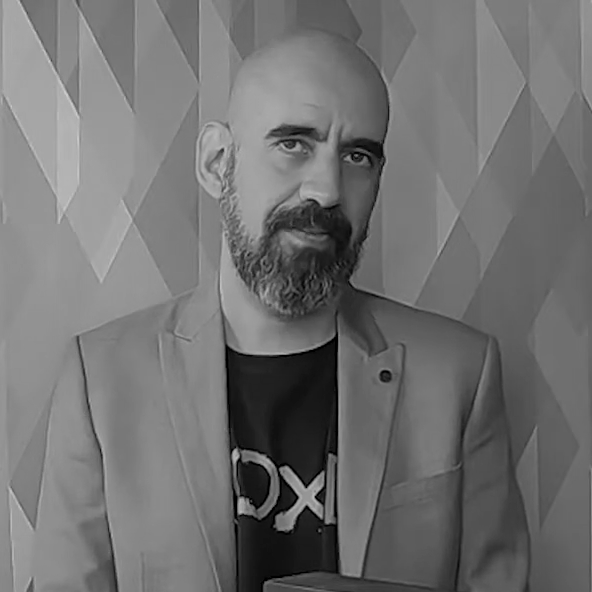 Black and white photo of a man with a beard and shaved head, wearing a blazer and a T-shirt, standing in front of a textured wall.
