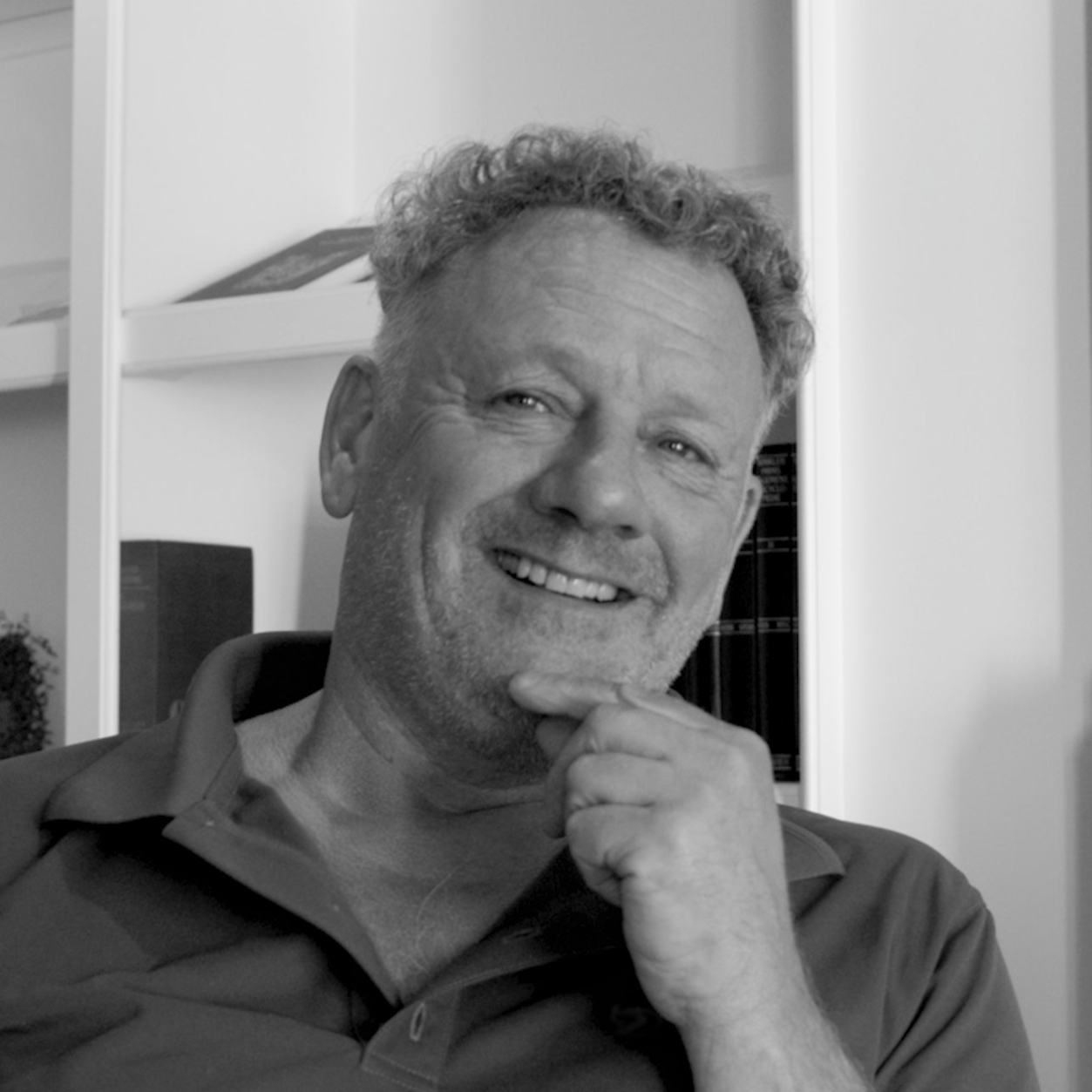A black-and-white photo of a smiling man with curly hair, resting his chin on his hand, in an indoor setting with shelves in the background.
