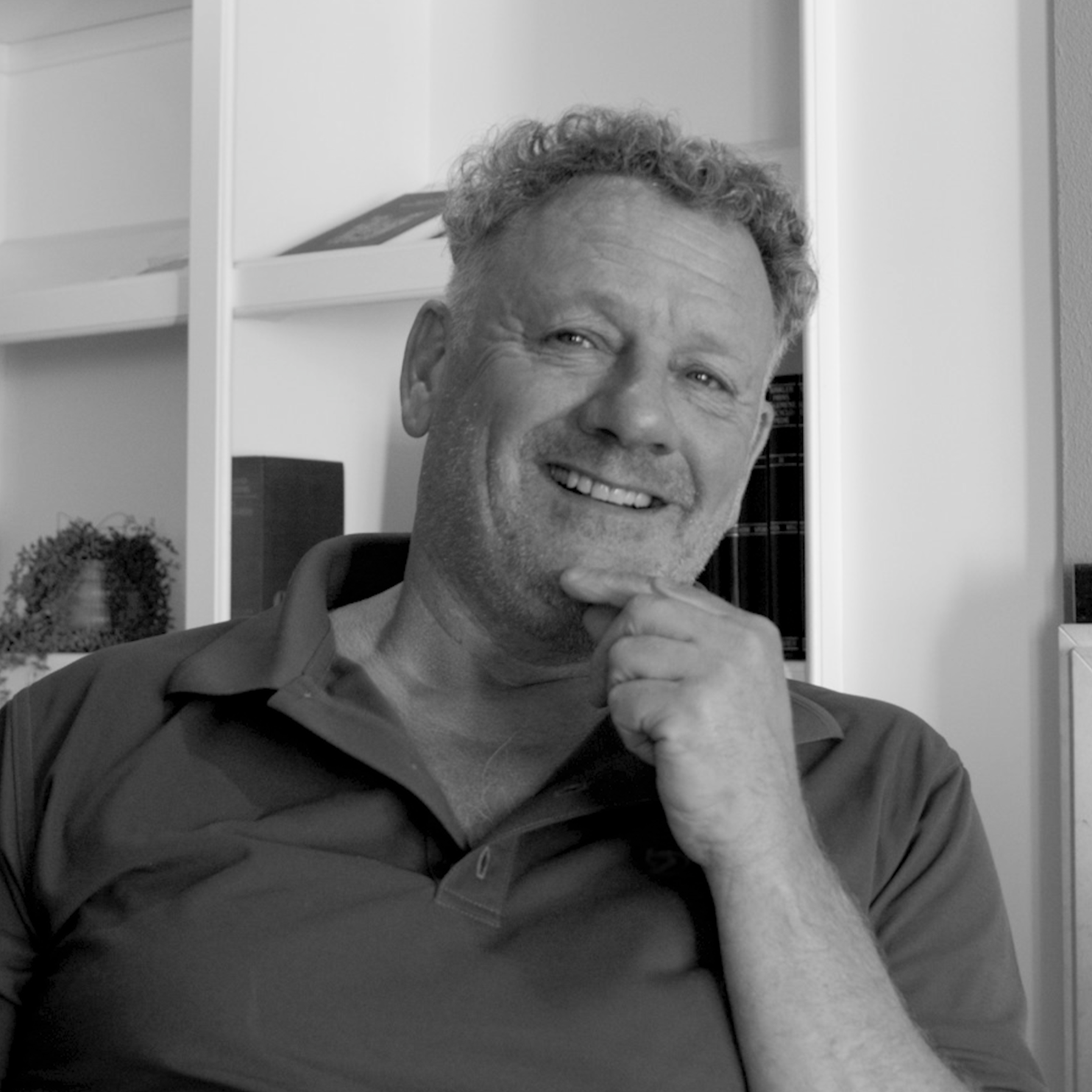 A smiling middle-aged man with curly hair and facial stubble, wearing a polo shirt, sitting indoors near a bookshelf, with one hand resting on his chin.