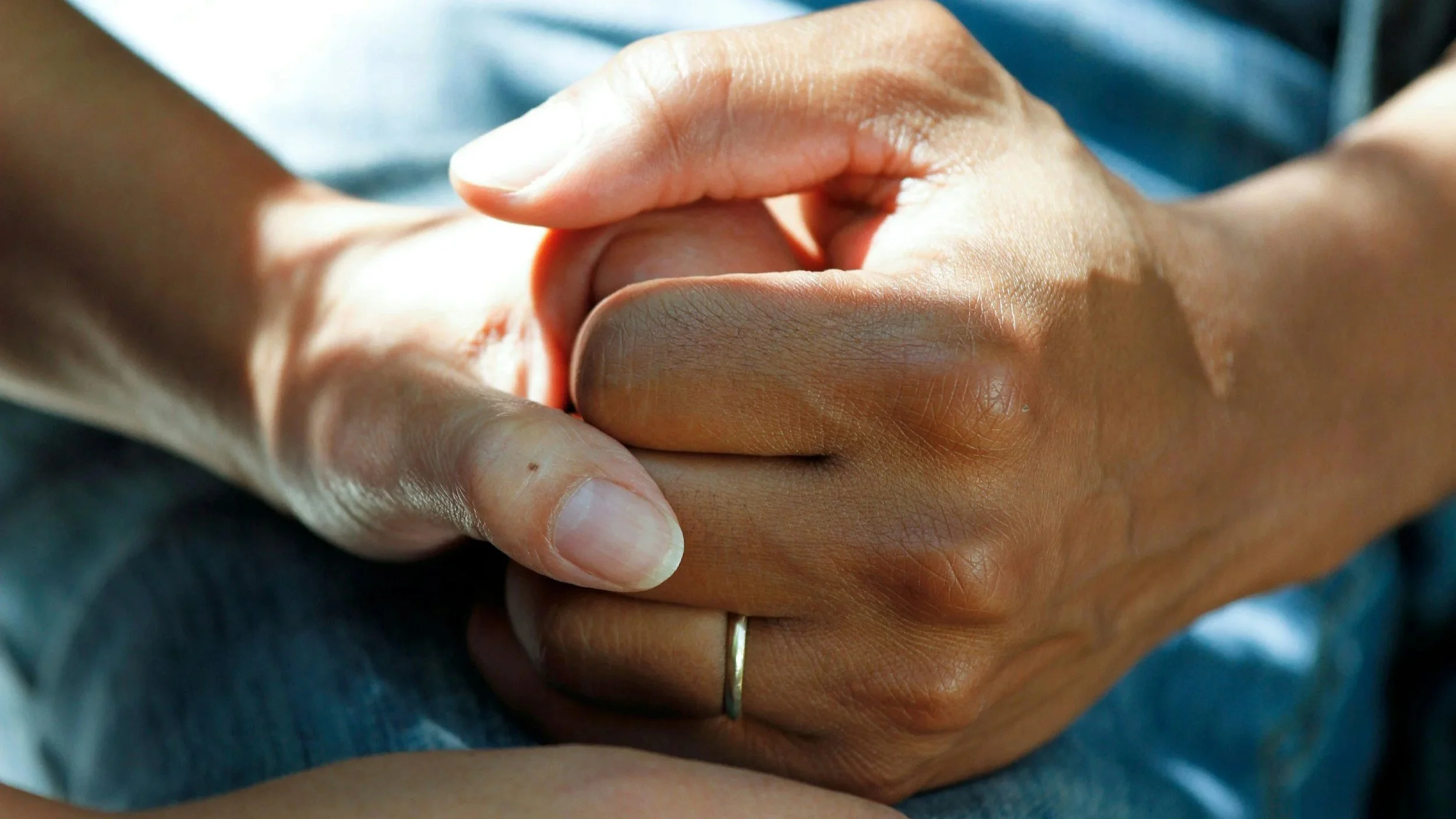 A close up of two people holding hands. The more masculine hand has a white gold wedding band on their ring finger.