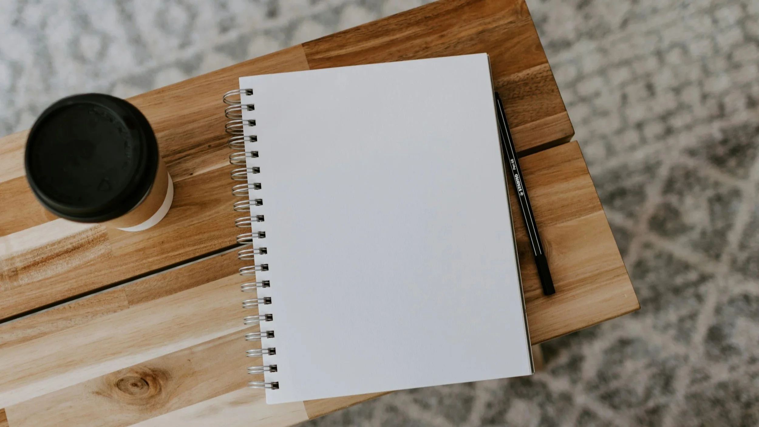 A spiral-bound notepad on a wooden table with a cup of tea and pen shot from above