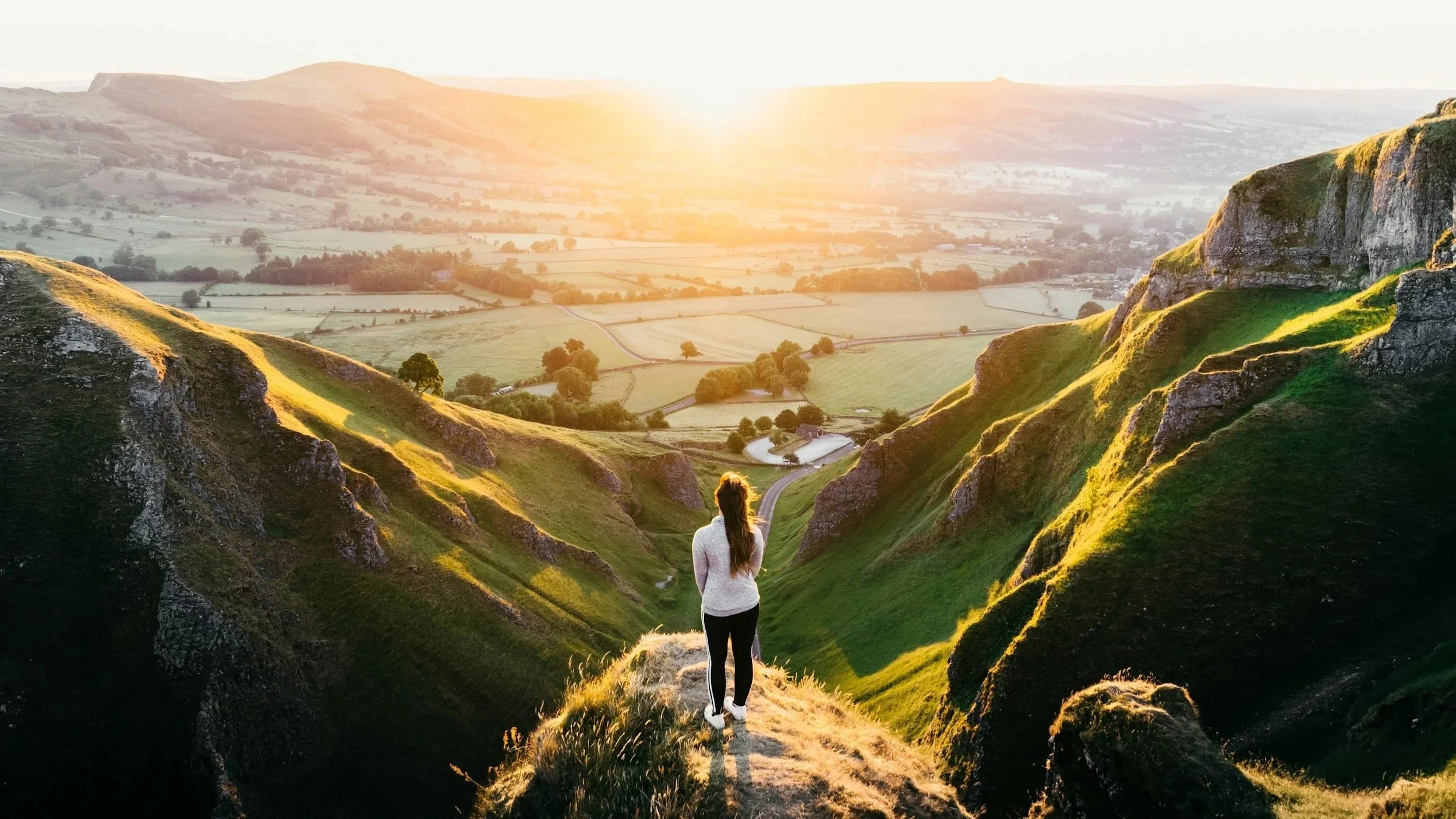 A woman standing on a rocky hill overlooking a valley at sunset, with rolled green hills, patches of farmland, and a winding road in the distance.