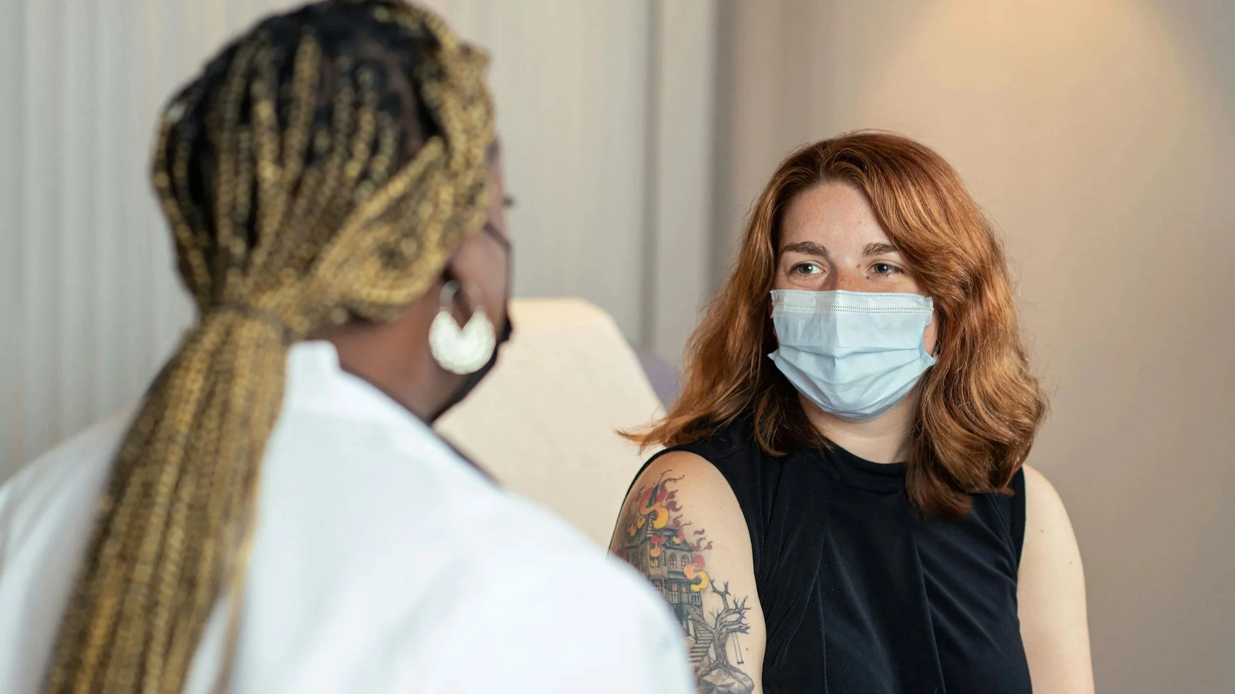 Two women, one black with long blonde braids, the other a white woman with copper hair face each other. They're wearing medical masks