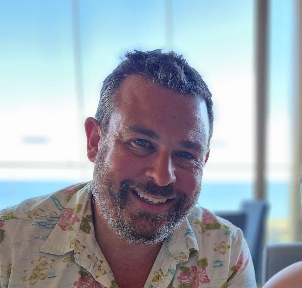 A smiling man with a beard and short hair, wearing a Hawaiian shirt, sitting indoors with natural light in the background.
