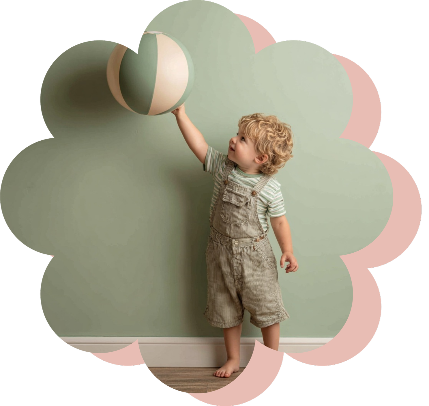 A young boy with curly blond hair standing barefoot against a light green wall, wearing beige overalls and a striped t-shirt, reaching out to touch a large, striped beach ball.