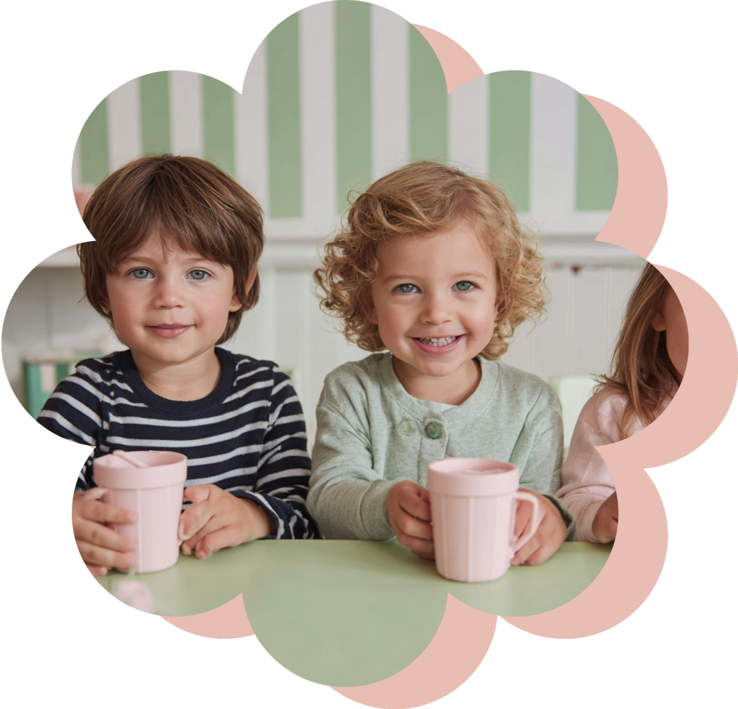 Two young children with curly and straight hair smiling and holding pink mugs, sitting at a table with a light green surface.