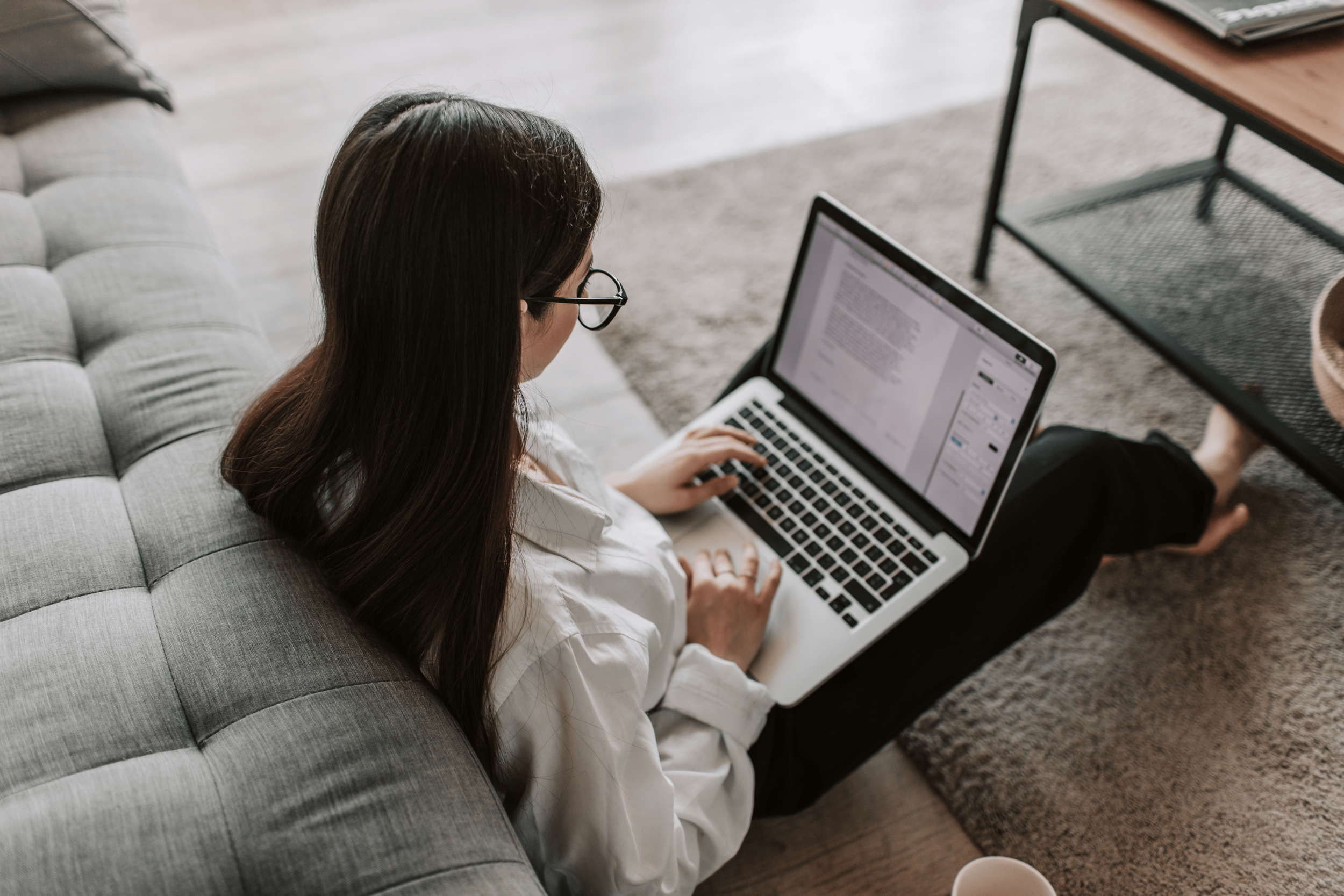 Woman with long dark hair, wearing glasses and a white shirt, sitting on a gray sofa working on a laptop with an open document, with a coffee mug nearby.