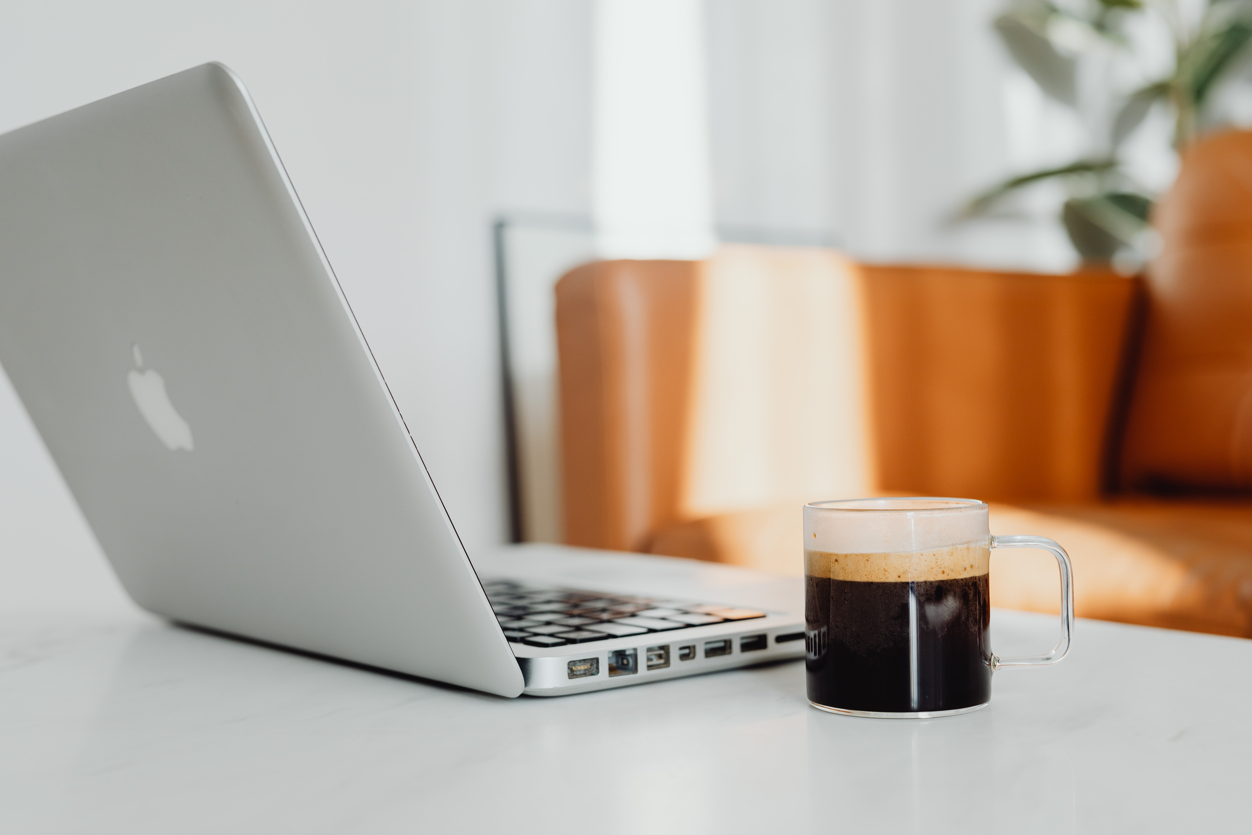 A silver MacBook laptop open on a white table with a clear glass mug of black coffee beside it, and a blurred background of a sofa and green plant.
