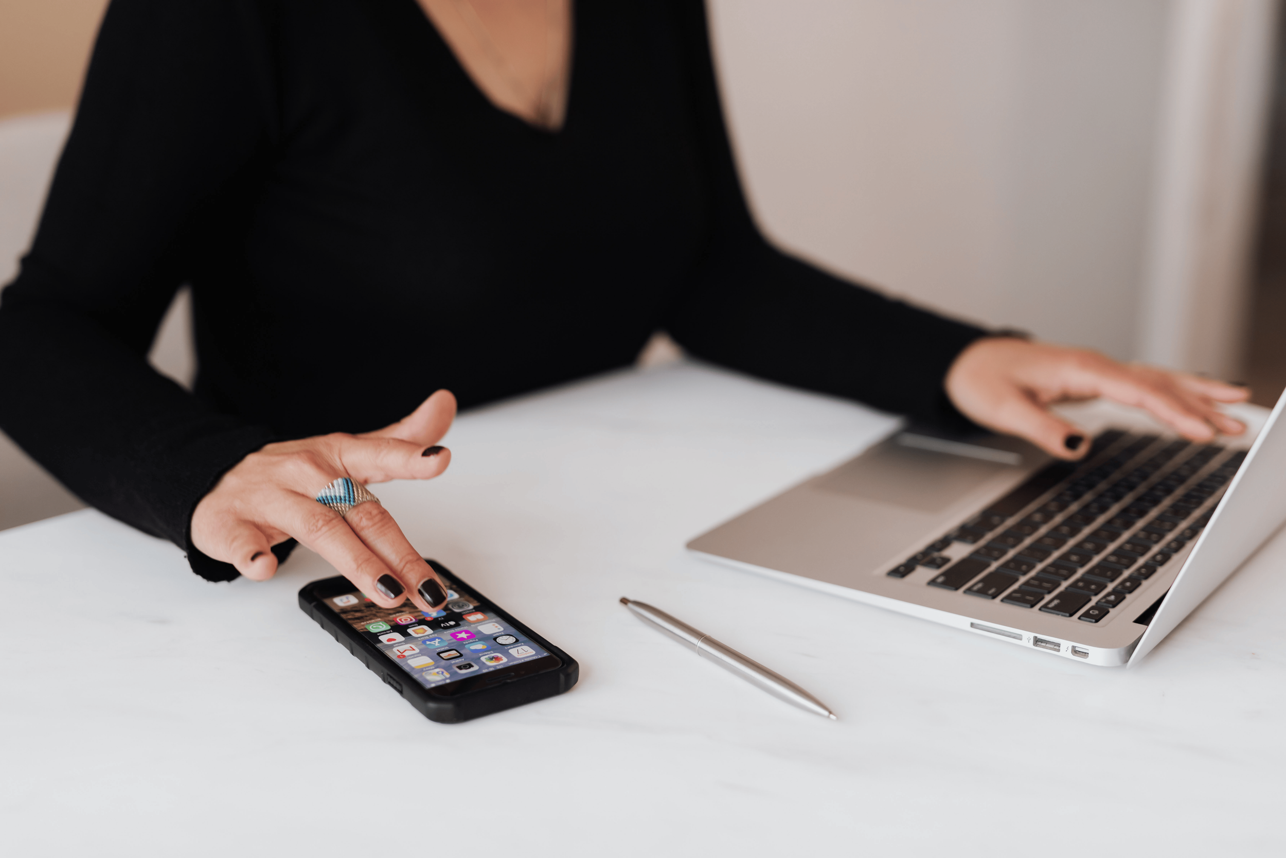 Person wearing a black top using a smartphone and a laptop on a white desk, with a silver pen placed nearby.