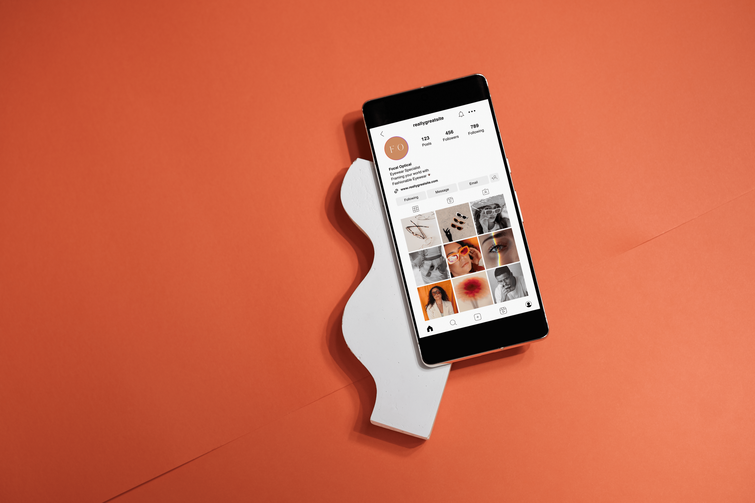 A smartphone displaying an Instagram profile with a grid of photos, resting on a white hand-shaped stand on an orange background.