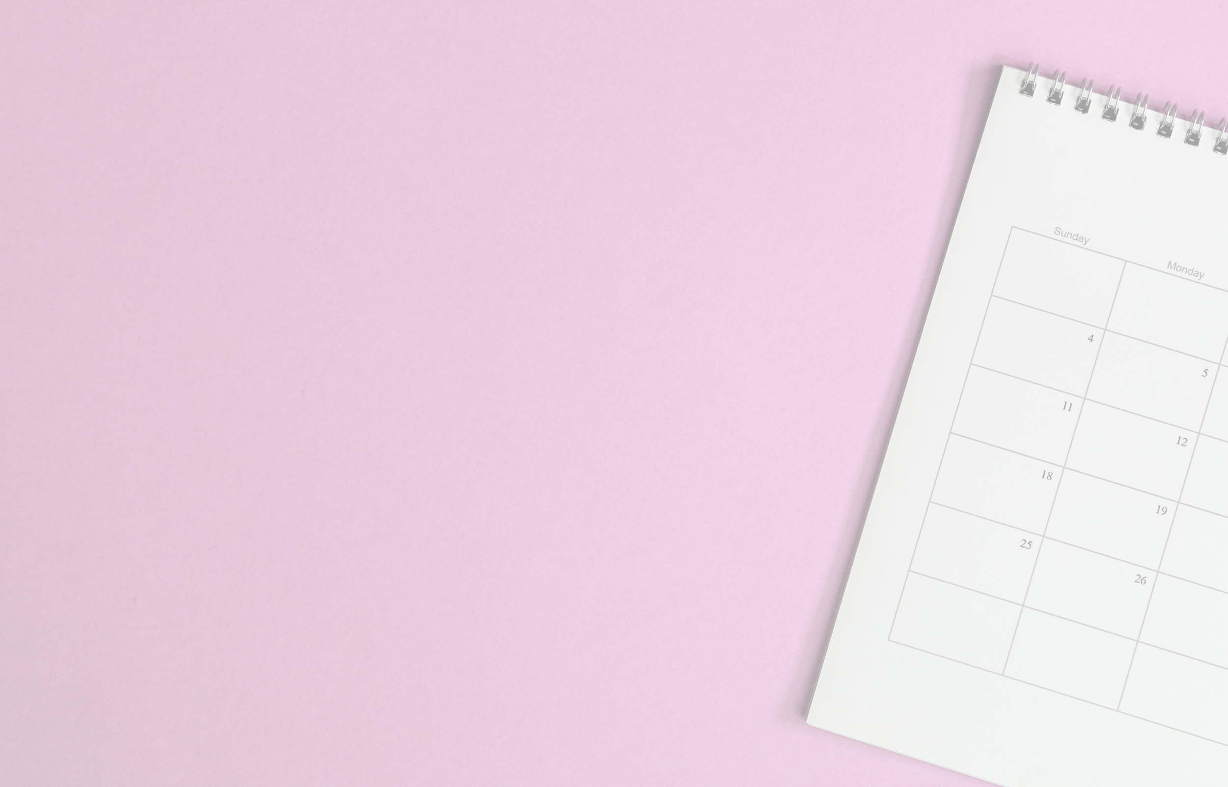 A white spiral-bound calendar on a pink background, with the calendar showing dates for a month.