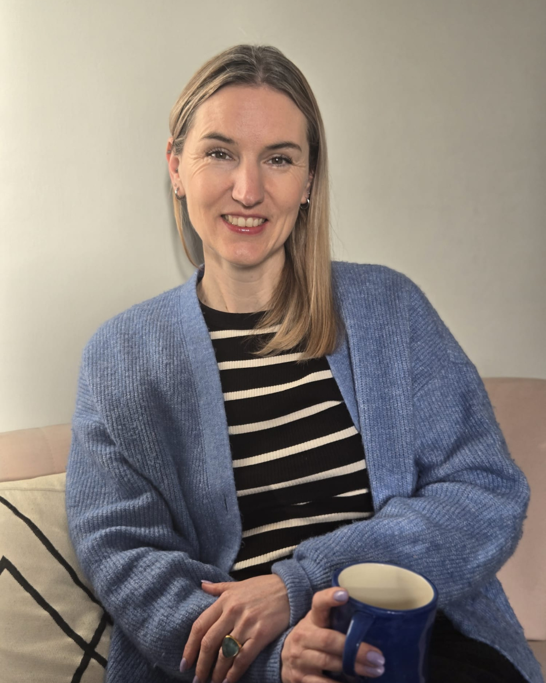 A woman with shoulder-length blonde hair, wearing a blue cardigan over a black and white striped shirt, smiling and holding a blue mug, sitting on a beige sofa with a cream-colored pillow.