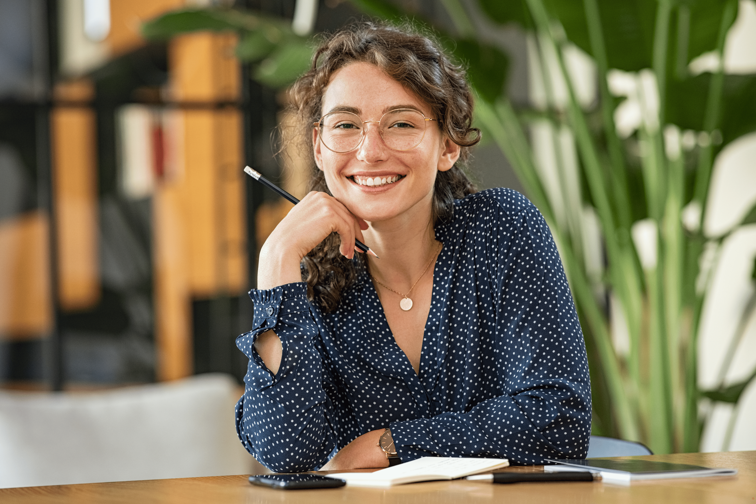 A woman with curly brown hair, glasses, and a polka-dot blouse smiling while sitting at a desk with a notebook, pen, and phone in front of her. Green plants and bookshelves are in the background.