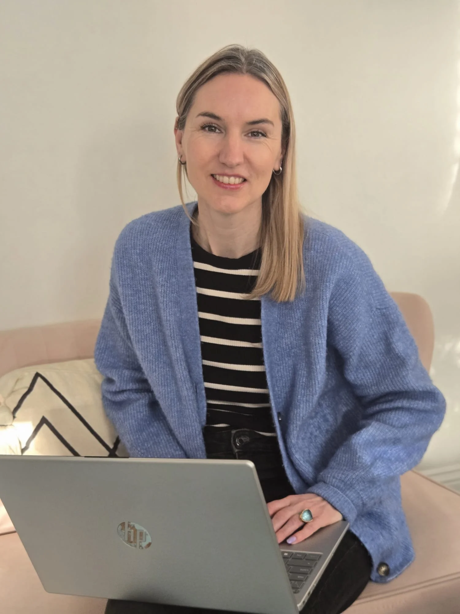 A young woman with shoulder-length blonde hair, wearing a black and white striped shirt and a blue cardigan, is sitting on a beige couch with a laptop on her lap, smiling at the camera.