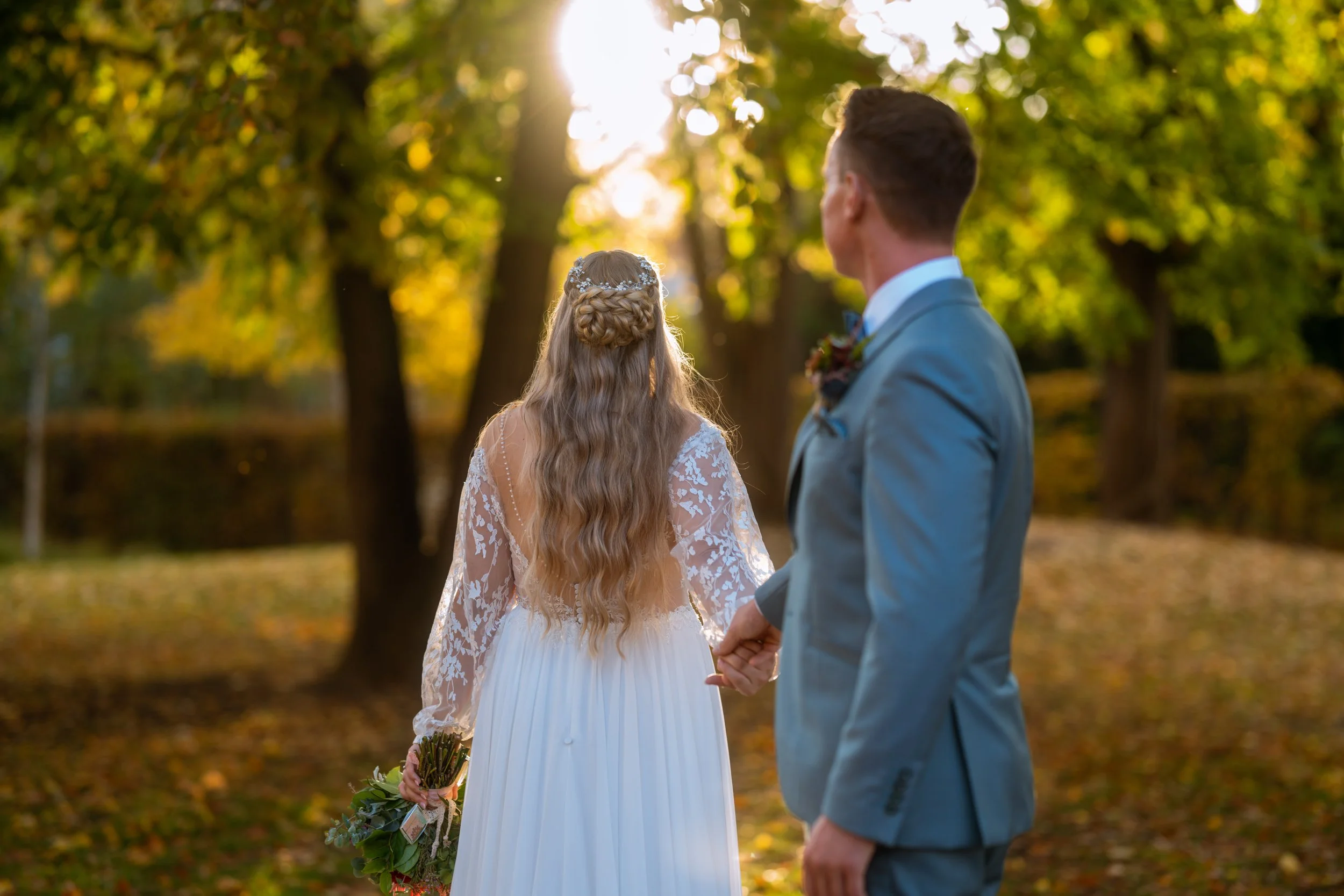 Braut und Bräutigam halten Händchen im Park bei Sonnenuntergang an einem Herbsttag, umgeben von gelb und grün gefärbten Bäumen, bei einer Hochzeitszeremonie.
