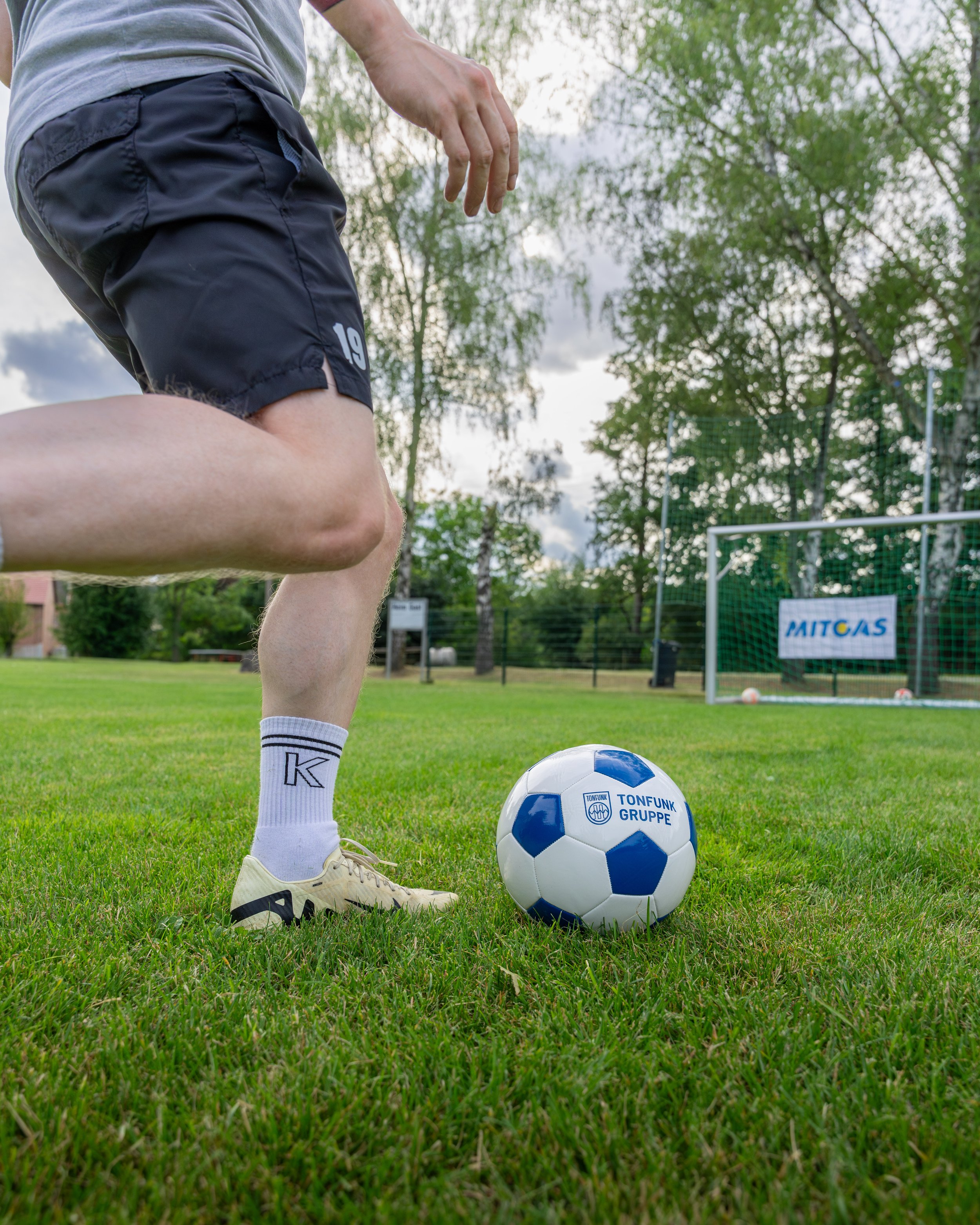 Fußballspieler steht auf dem Rasen vor einem Fußball mit der englischen Aufschrift 'TONFUNK GRUPPE' und bereitet sich auf einen Schuss vor, mit Tor und Banden im Hintergrund.