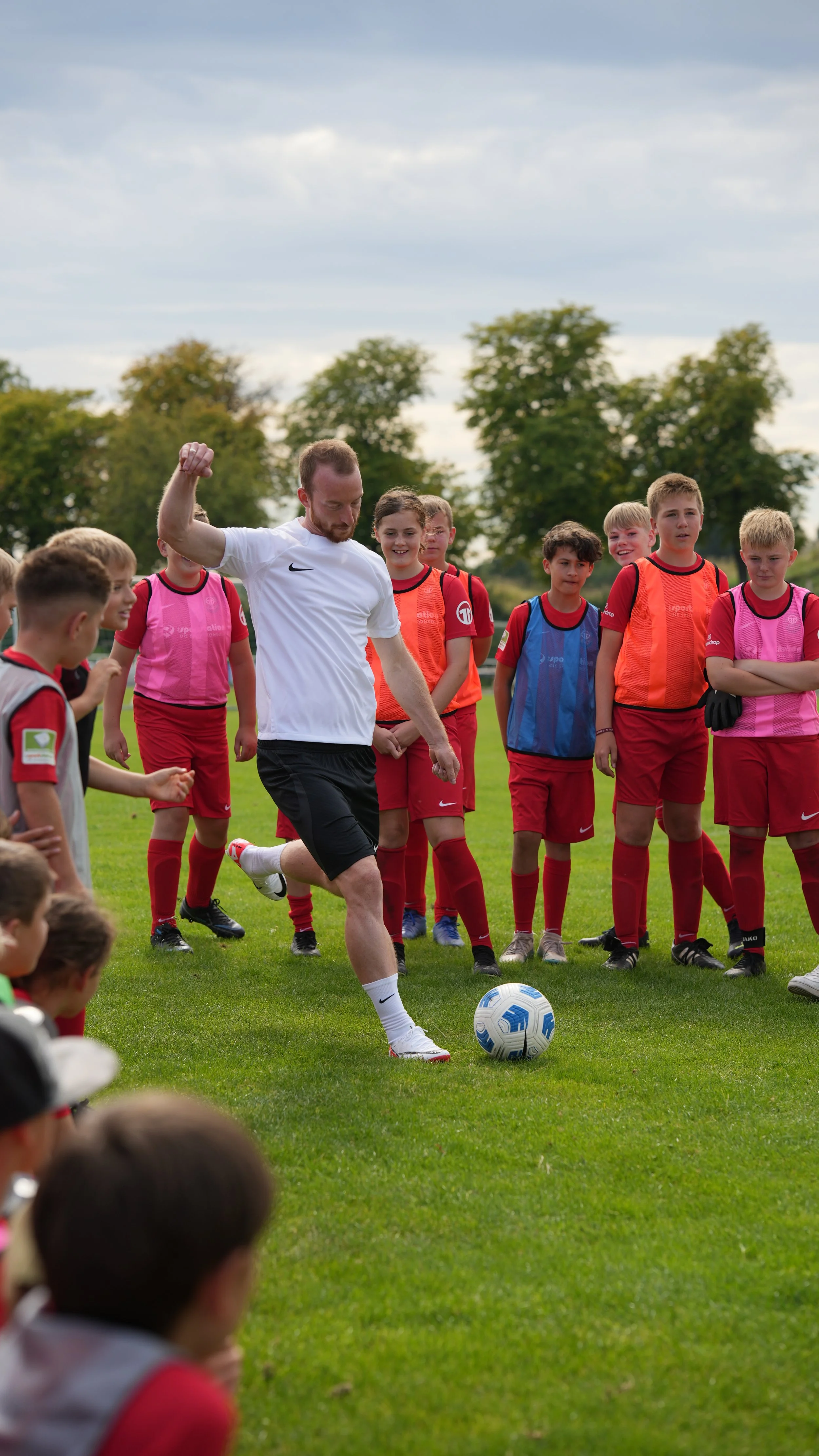 Ein männlicher Trainer oder Spieler übt mit jungen Fußballspielern auf einem Sportplatz, indem er vor ihnen einen Ball schießt.