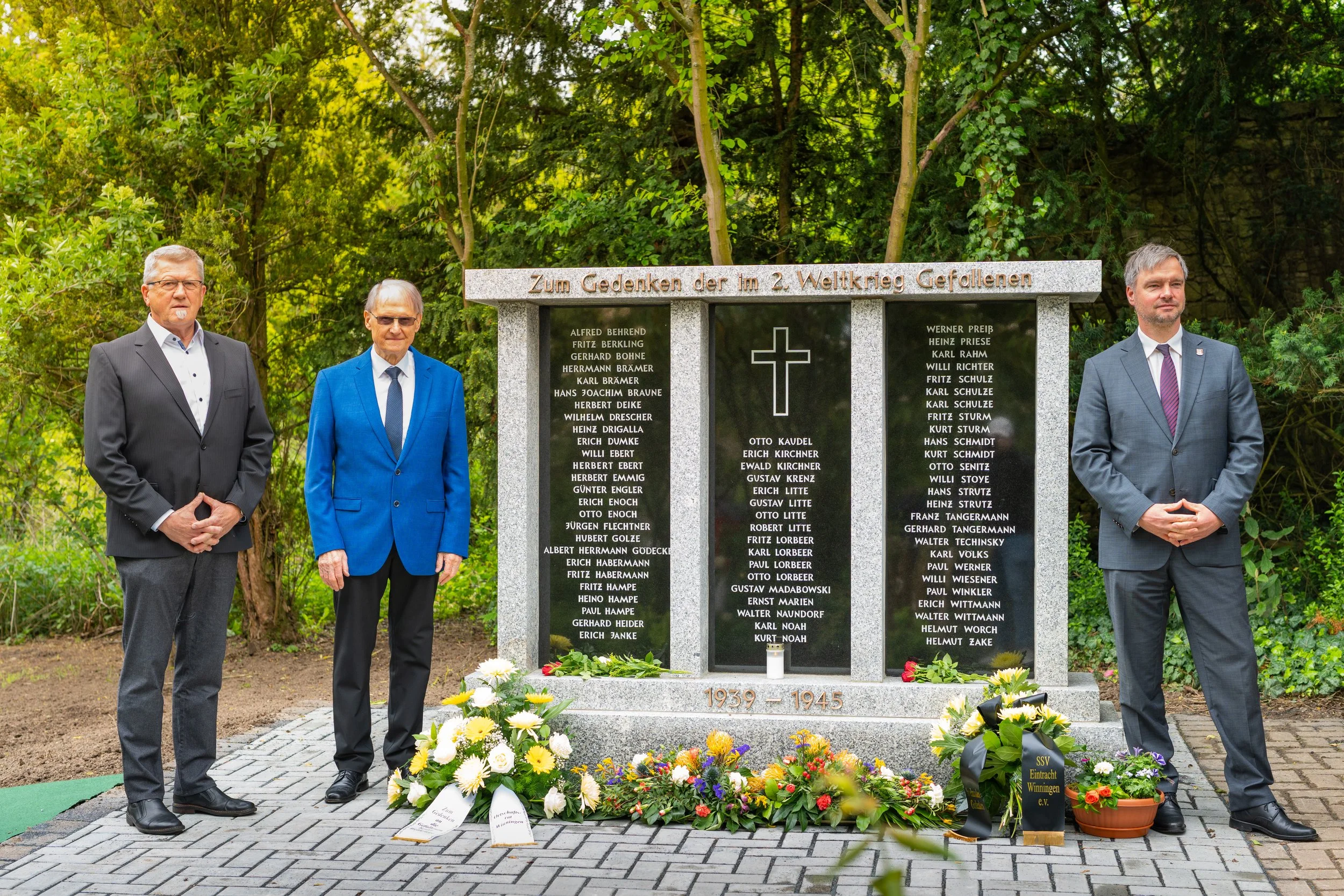Denkmal mit Namen gefallener Soldaten im Zweiten Weltkrieg, flankiert von drei Männern in Anzügen, im Hintergrund Bäume und Pflanzen, Blumen und Kerzen am Denkmal