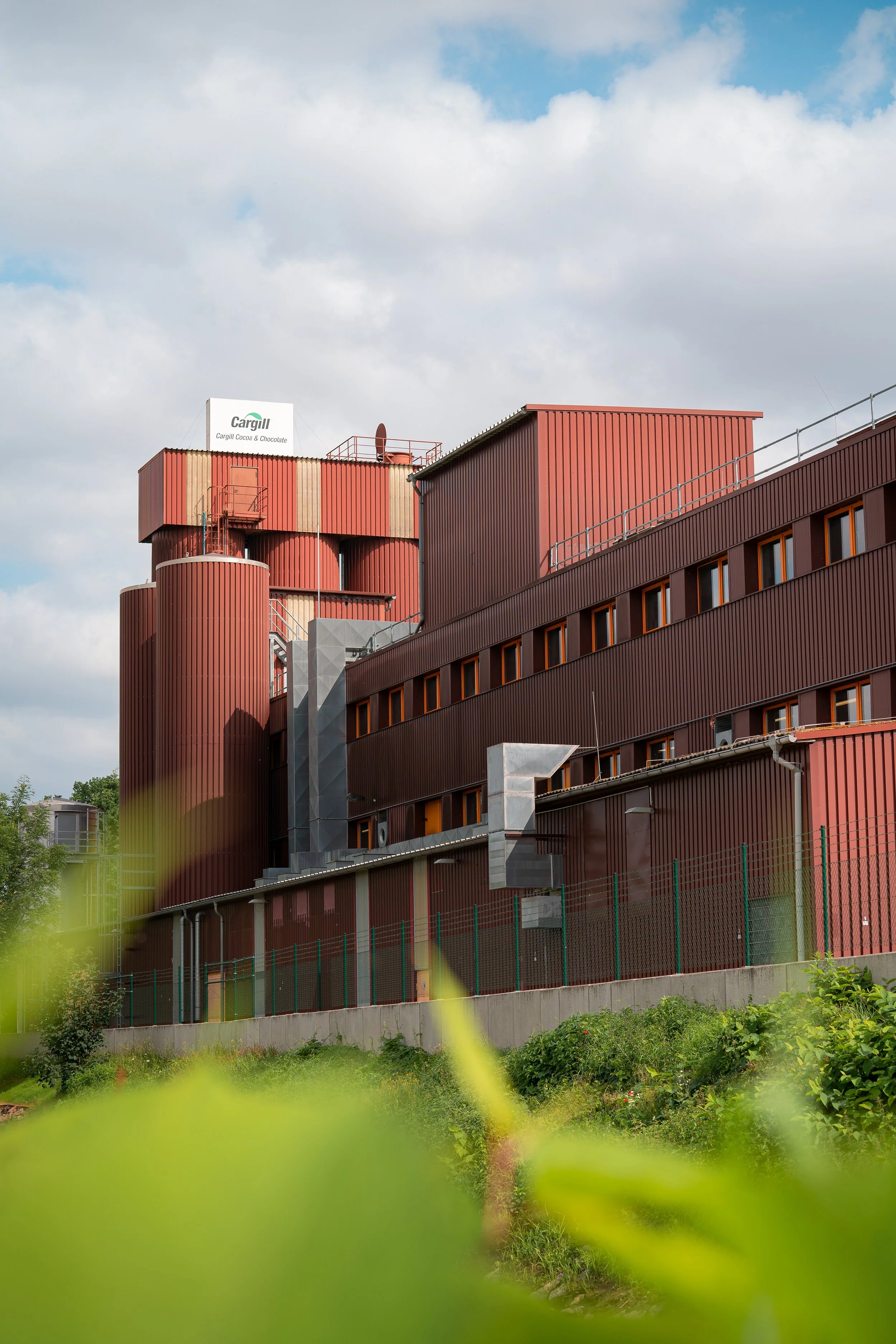 Ein modernes, rotes Fabrikgebäude mit runden Silos und einem angularen oberen Abschnitt. Das Gebäude hat mehrere Fenster und ist von einem grünen Garten mit Büschen und Pflanzen umgeben. Der Himmel ist bewölkt mit einigen blauen Flecken.