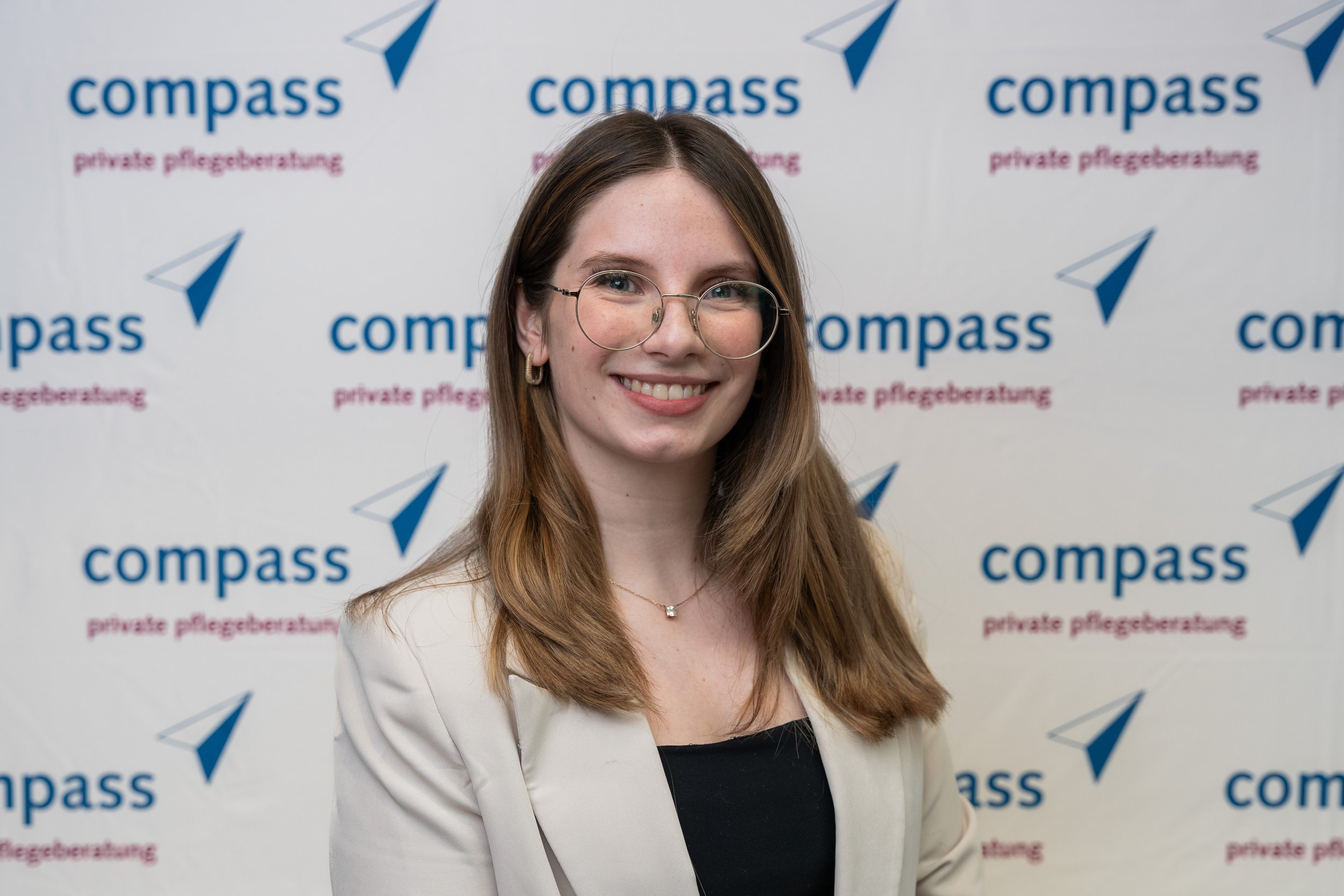 Junge Frau mit braunen Haaren, Brille und freundlichem Lächeln vor einem Hintergrund mit dem Logo und der Aufschrift "compass" und "private Pflegeberatung" auf weißem Hintergrund.