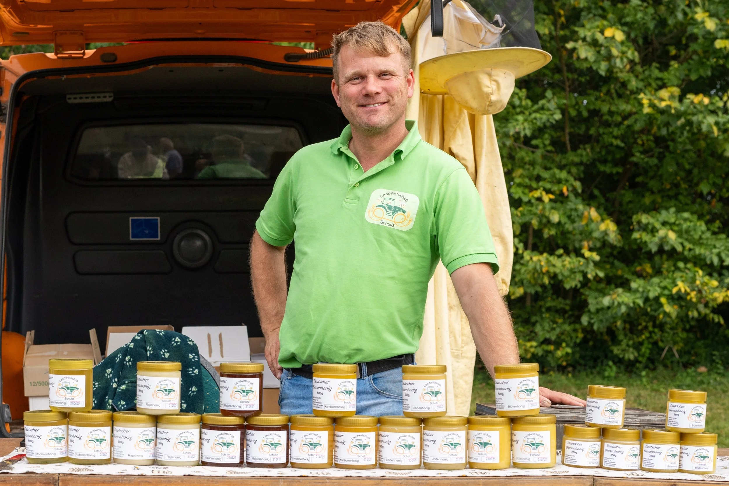 Ein Mann steht bei einem Verkaufsstand mit Honiggläsern, trägt ein grünes T-Shirt mit einem Landwirtschaftslogo, hinter ihm ein Auto mit geöffnetem Kofferraum. Es ist im Freien, umgeben von Bäumen.