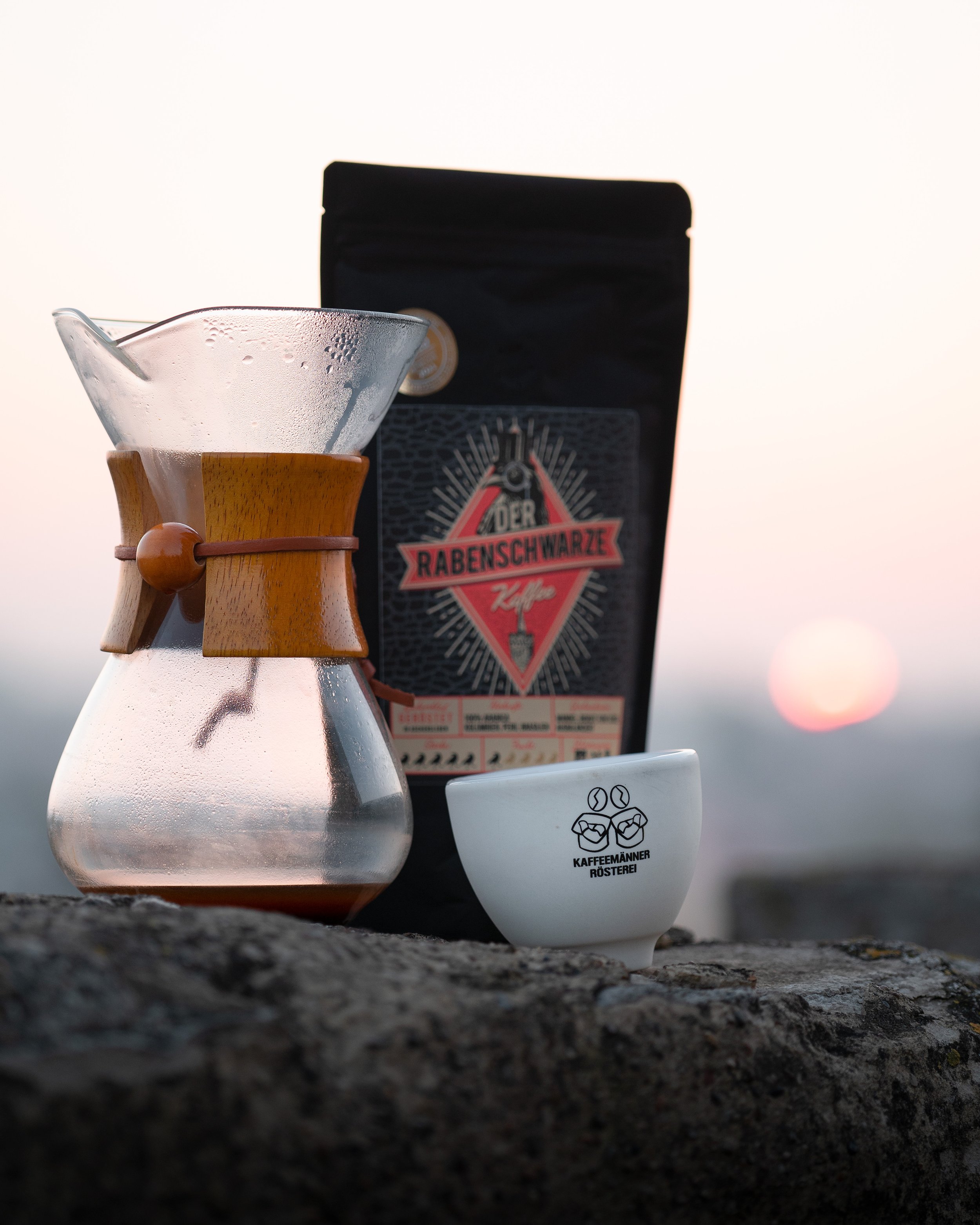 Eine Kaffeekanne, eine Tasse mit einem Logo der Kaffeerösterei Rabenschwarze, eine Kaffeemaschine, die im Hintergrund steht, und eine Tasse mit dem Logo der Kaffeemänner Rösterei, alles auf einer steinigen Oberfläche bei Sonnenuntergang.