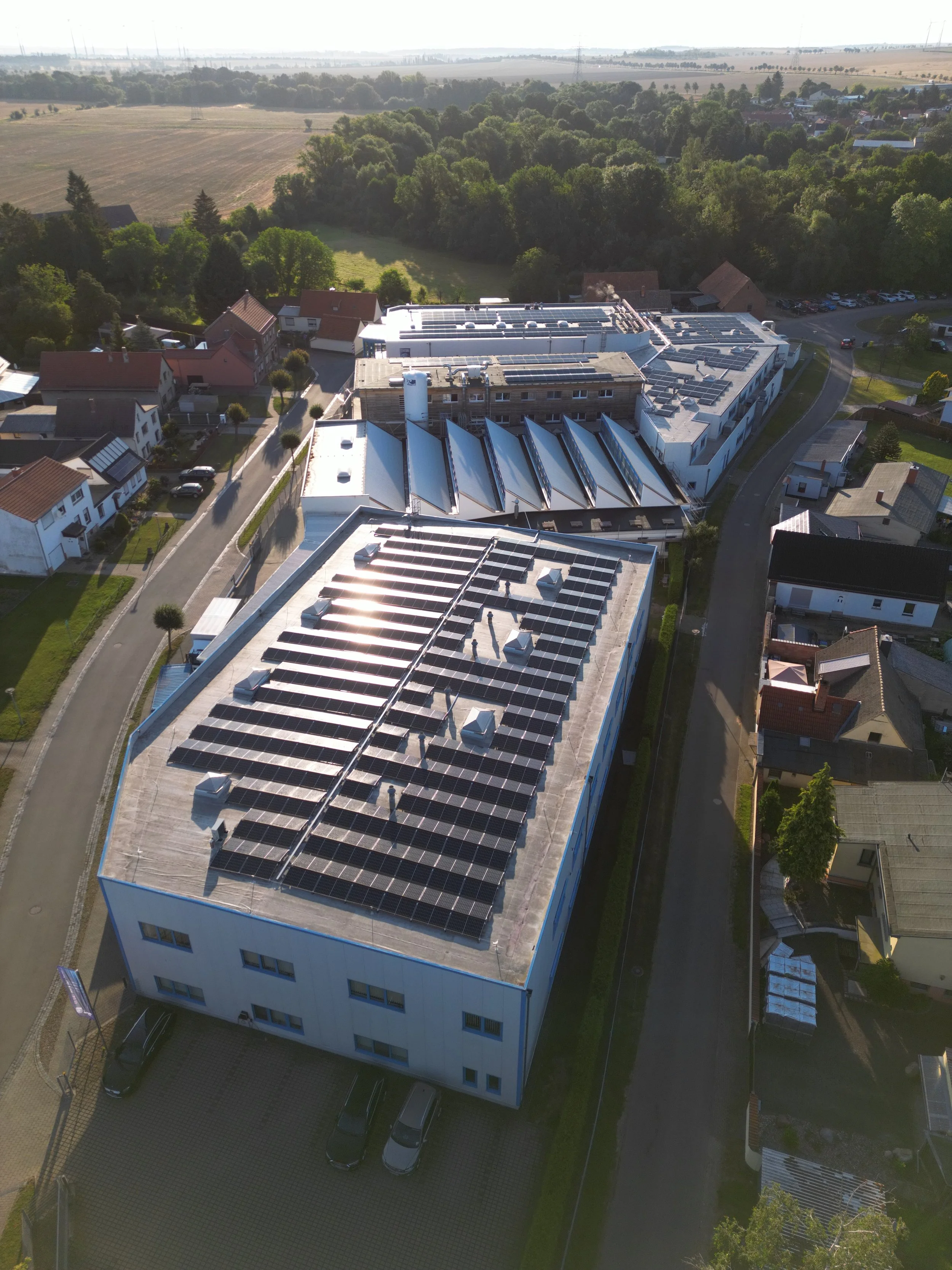 Ein Gebäude mit Solaranlagen auf dem Dach, umgeben von einem Wohngebiet mit Einfamilienhäusern und viel Grün im Hintergrund.