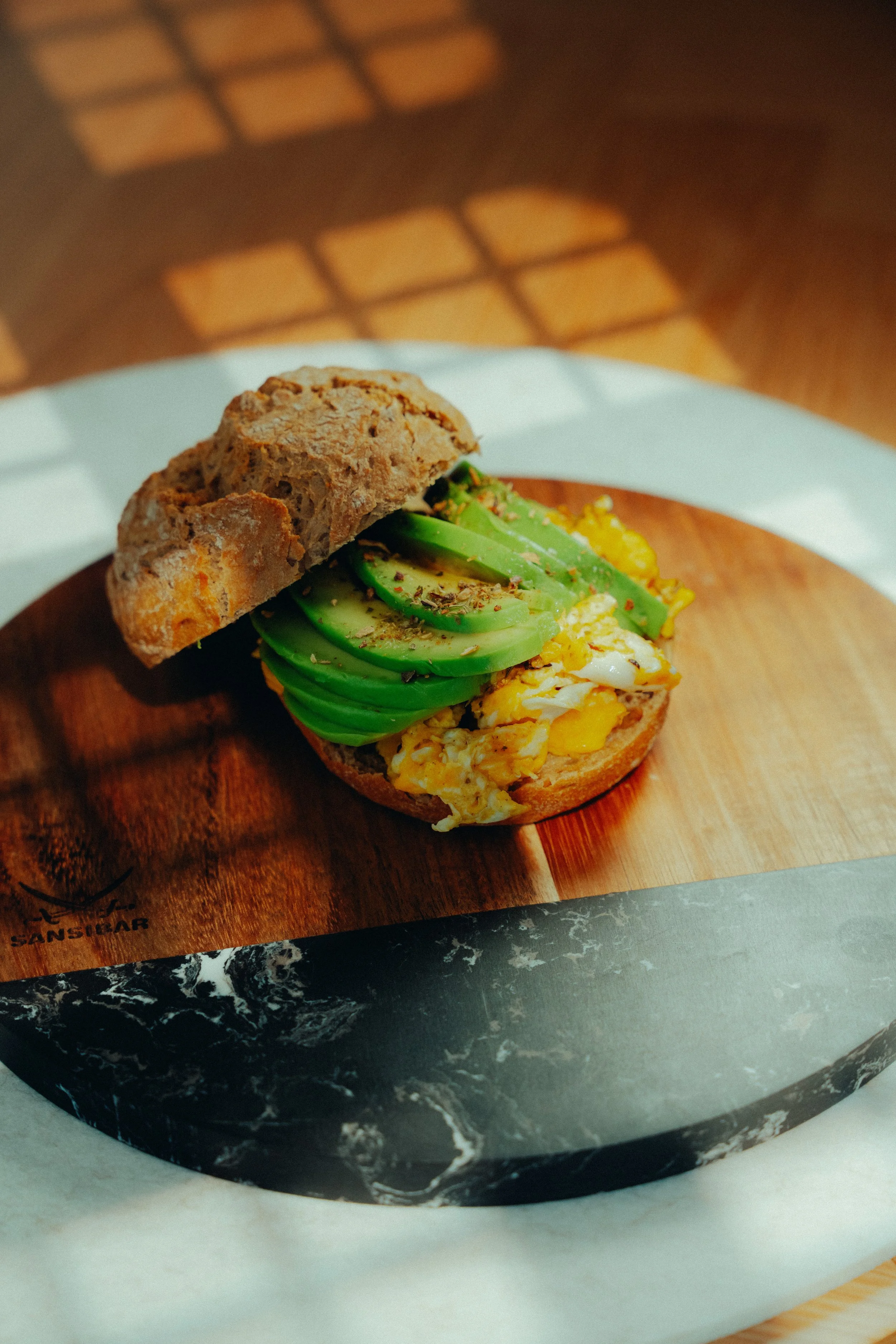 Ein belegtes Brötchen mit Avocado, Rührei und Brot auf einem Holzbrett, vor einer schwarzen Marmor-Plate